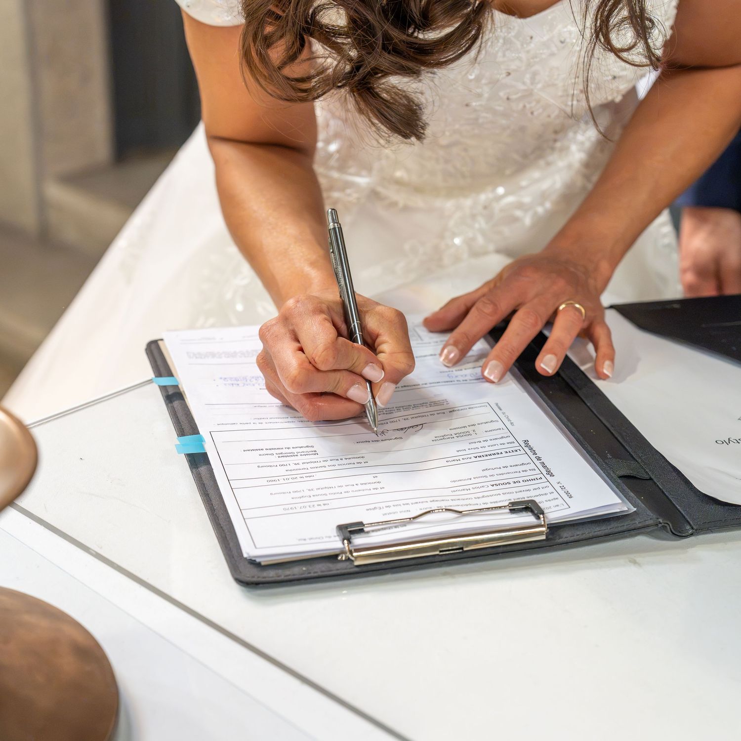 Bride in white lace dress signing marriage certificate on clipboard with pen at wedding ceremony.