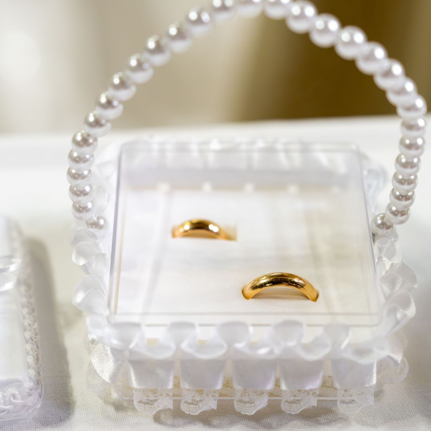 Two gold wedding bands displayed in a clear acrylic box with pearl handle and lace trim details.