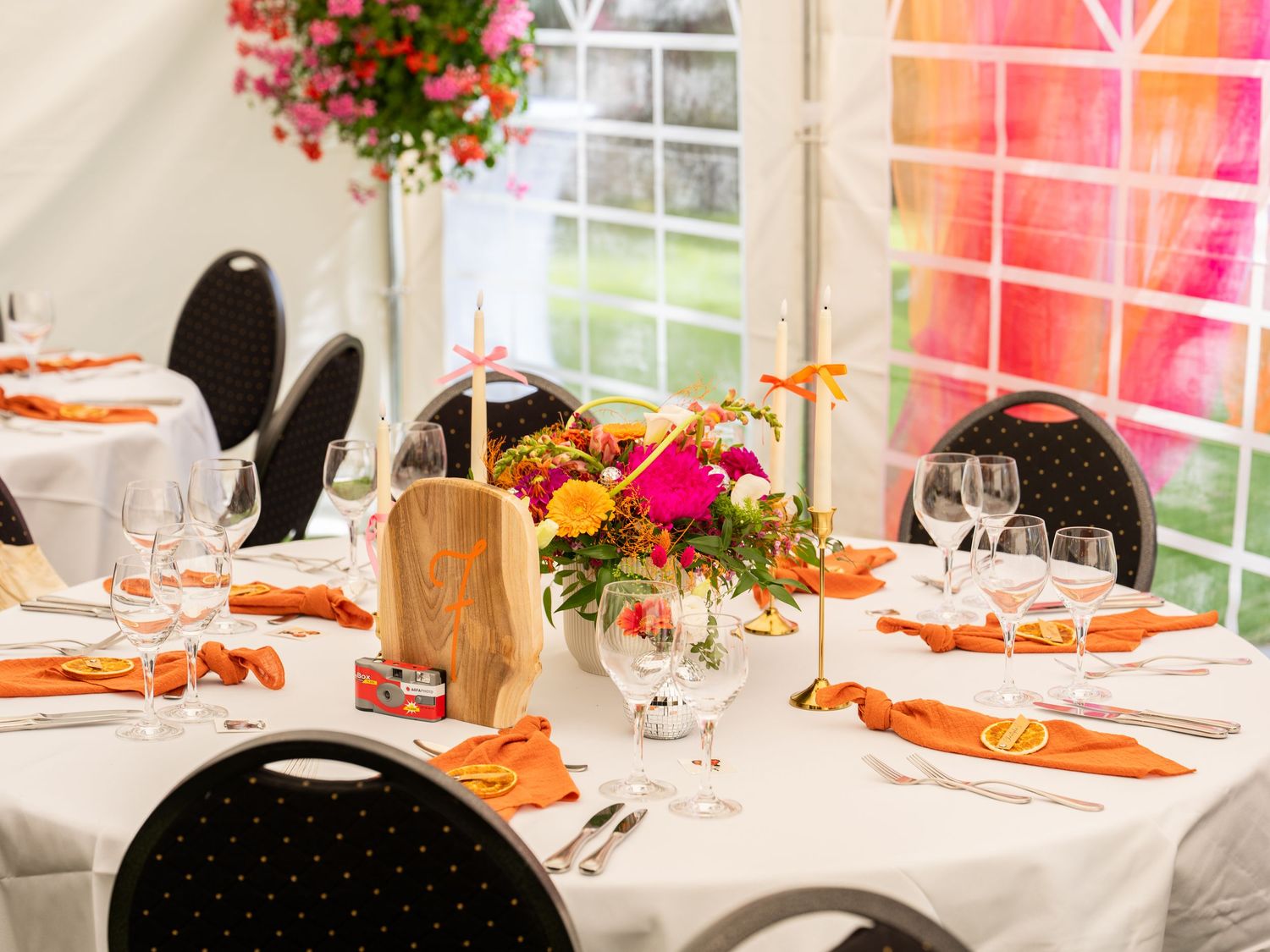 Elegant wedding reception table with orange napkins, floral centerpiece, and crystal glassware in a bright marquee tent.