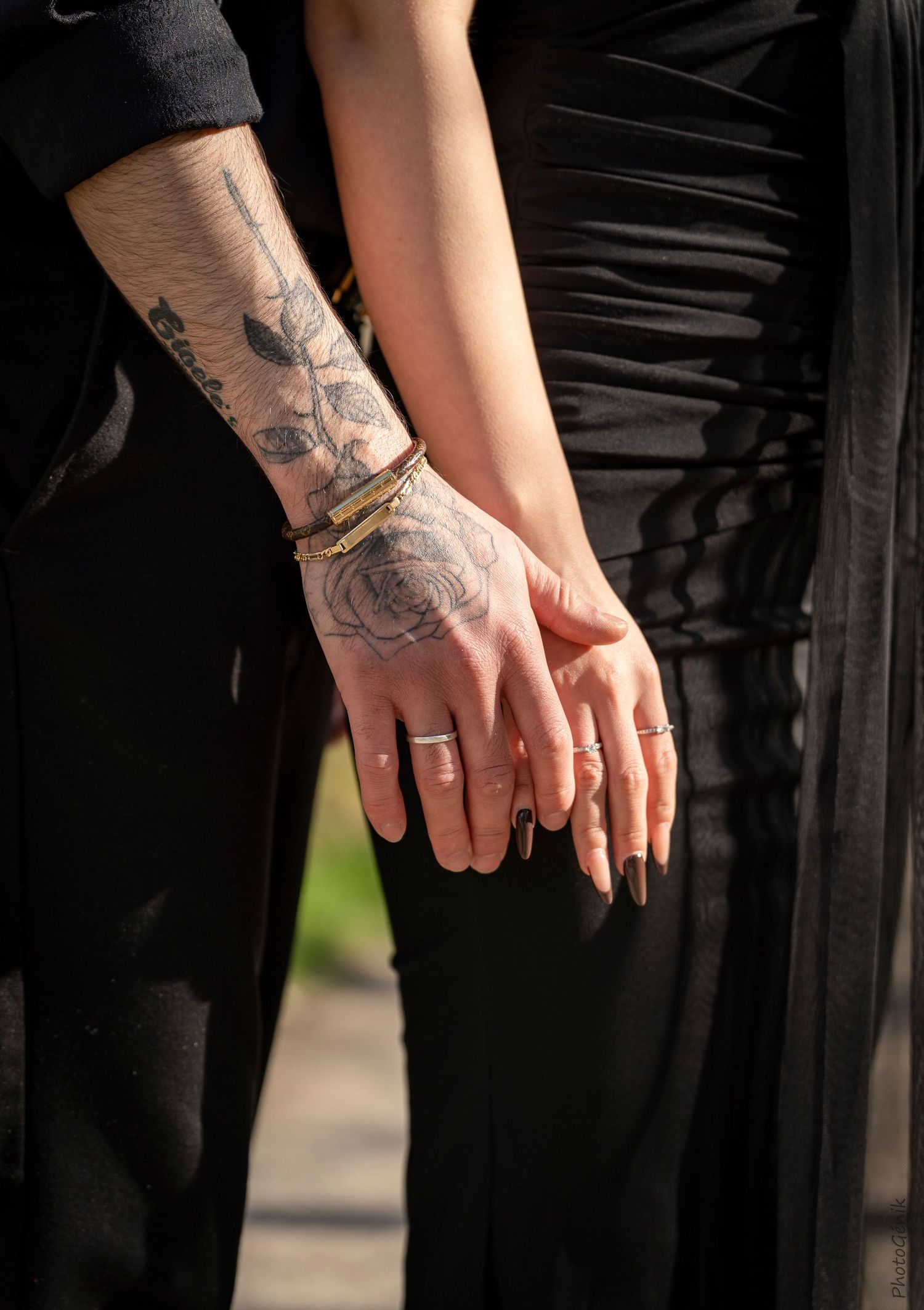 Two hands holding, one with rose tattoos and a gold bracelet, both wearing rings, dressed in black.