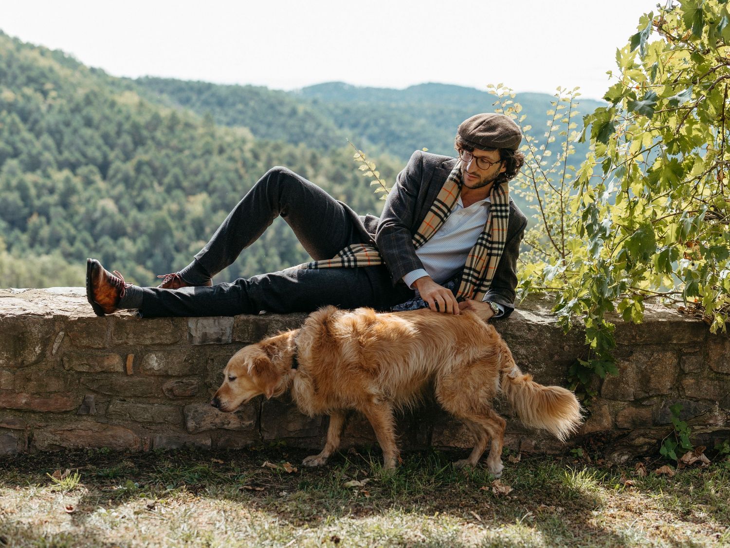 Casual man in plaid jacket relaxes on stone wall while petting a golden retriever in scenic landscape.