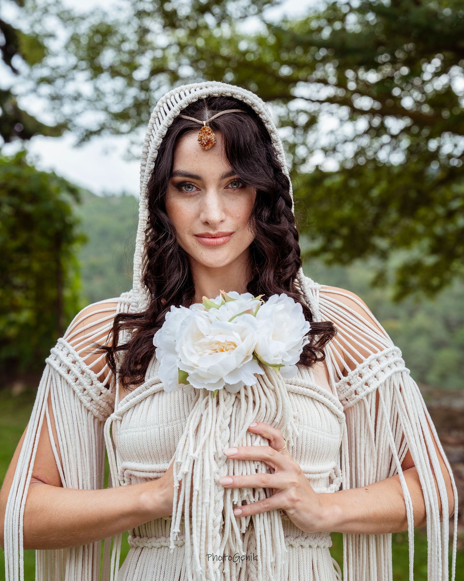 Bohemian bride in macrame hooded cape holding white floral bouquet with ornate gold headpiece outdoors.