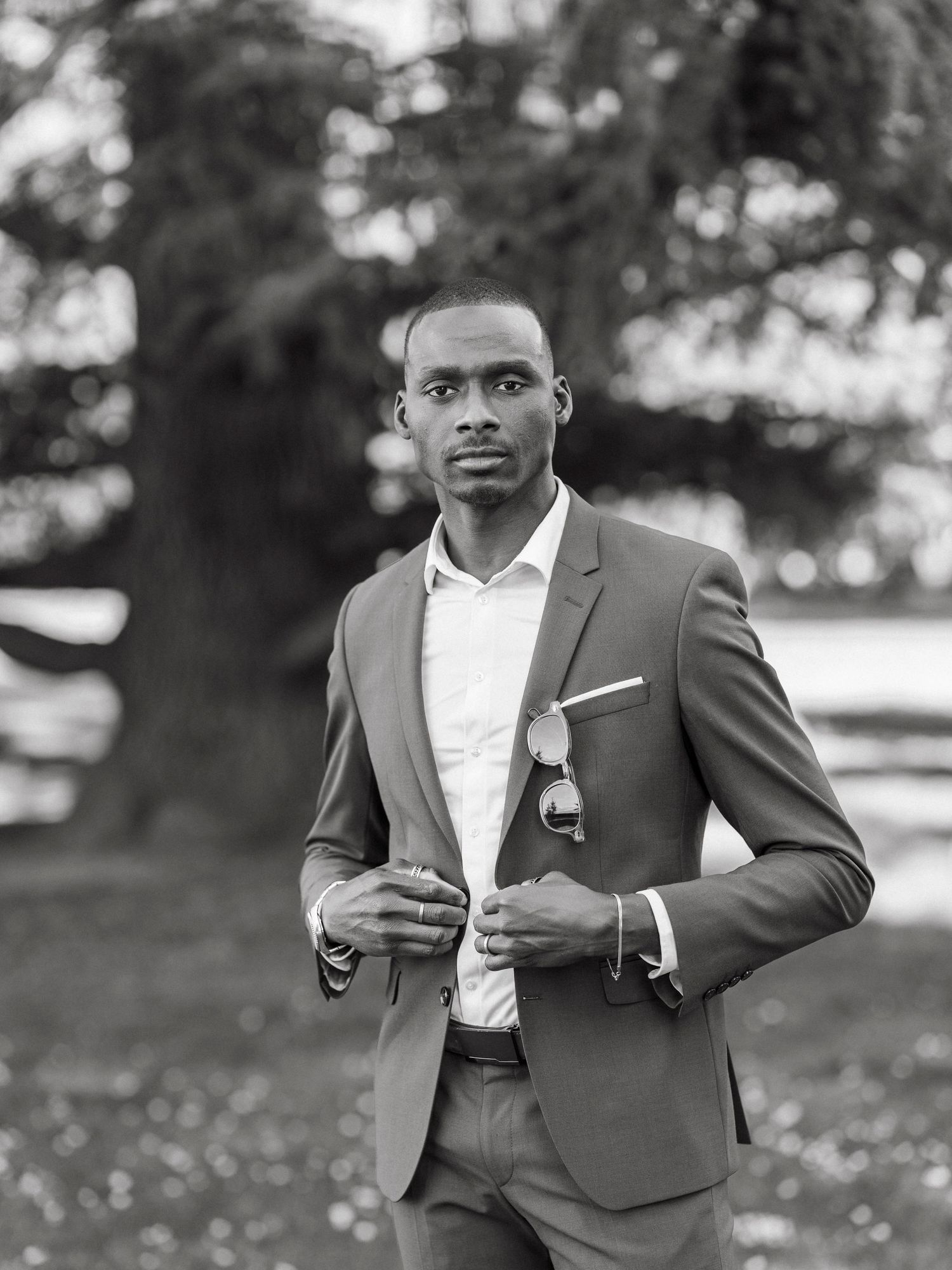 Stylish Black man in a tailored suit buttoning jacket outdoors in a park setting.