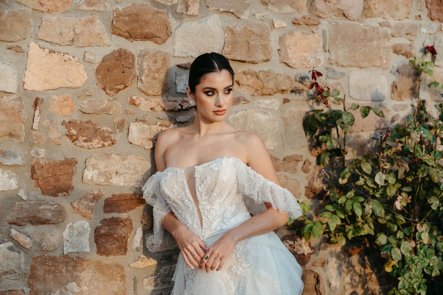 Bride in an off-shoulder lace ball gown poses gracefully against a rustic stone wall with ivy accents.