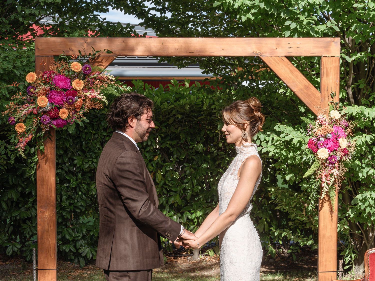 Couple exchanging vows under a wooden arch adorned with pink and orange floral arrangements at an outdoor wedding.