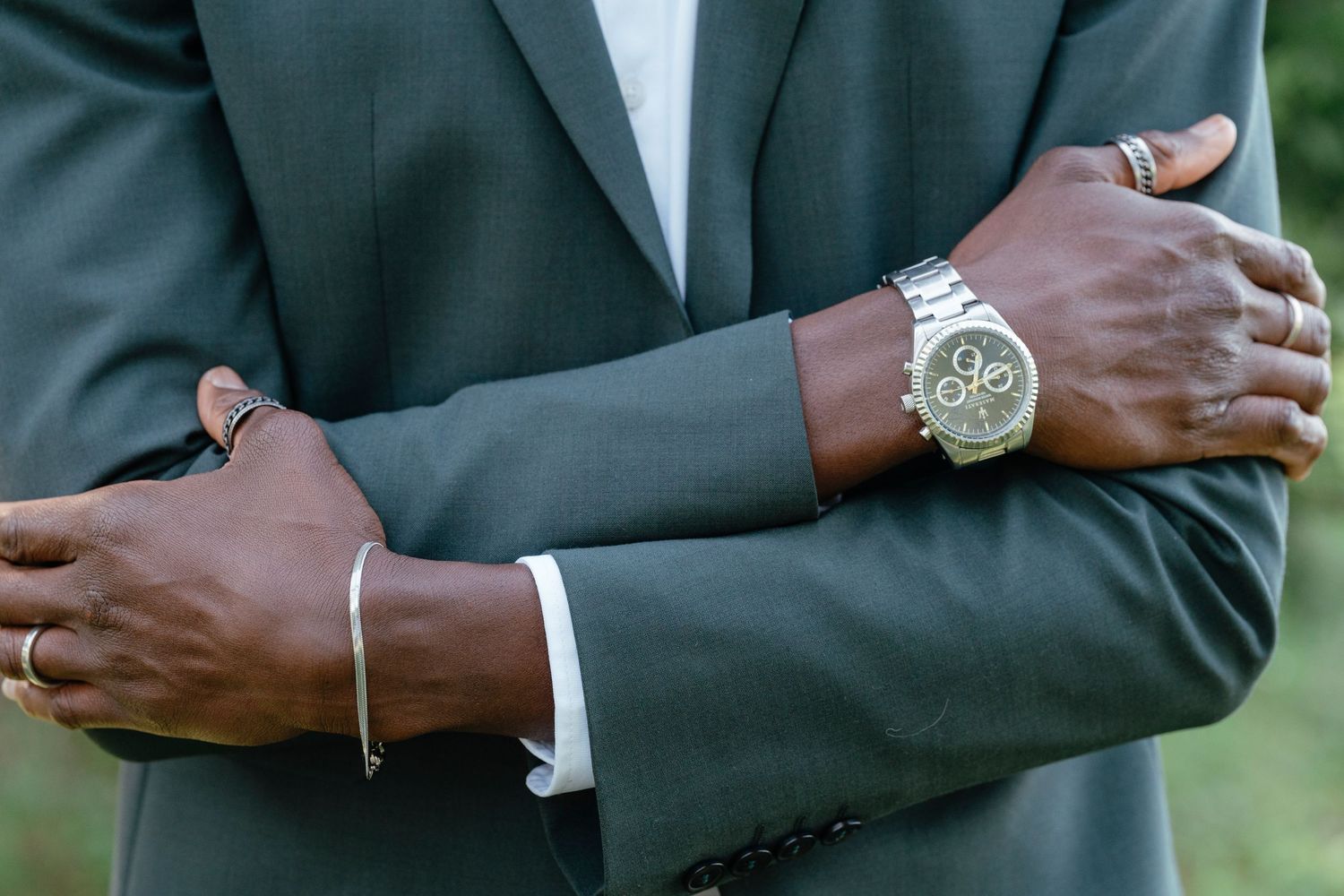 Elegant man in gray suit with crossed arms wearing a silver bracelet and luxury chronograph watch outdoors.