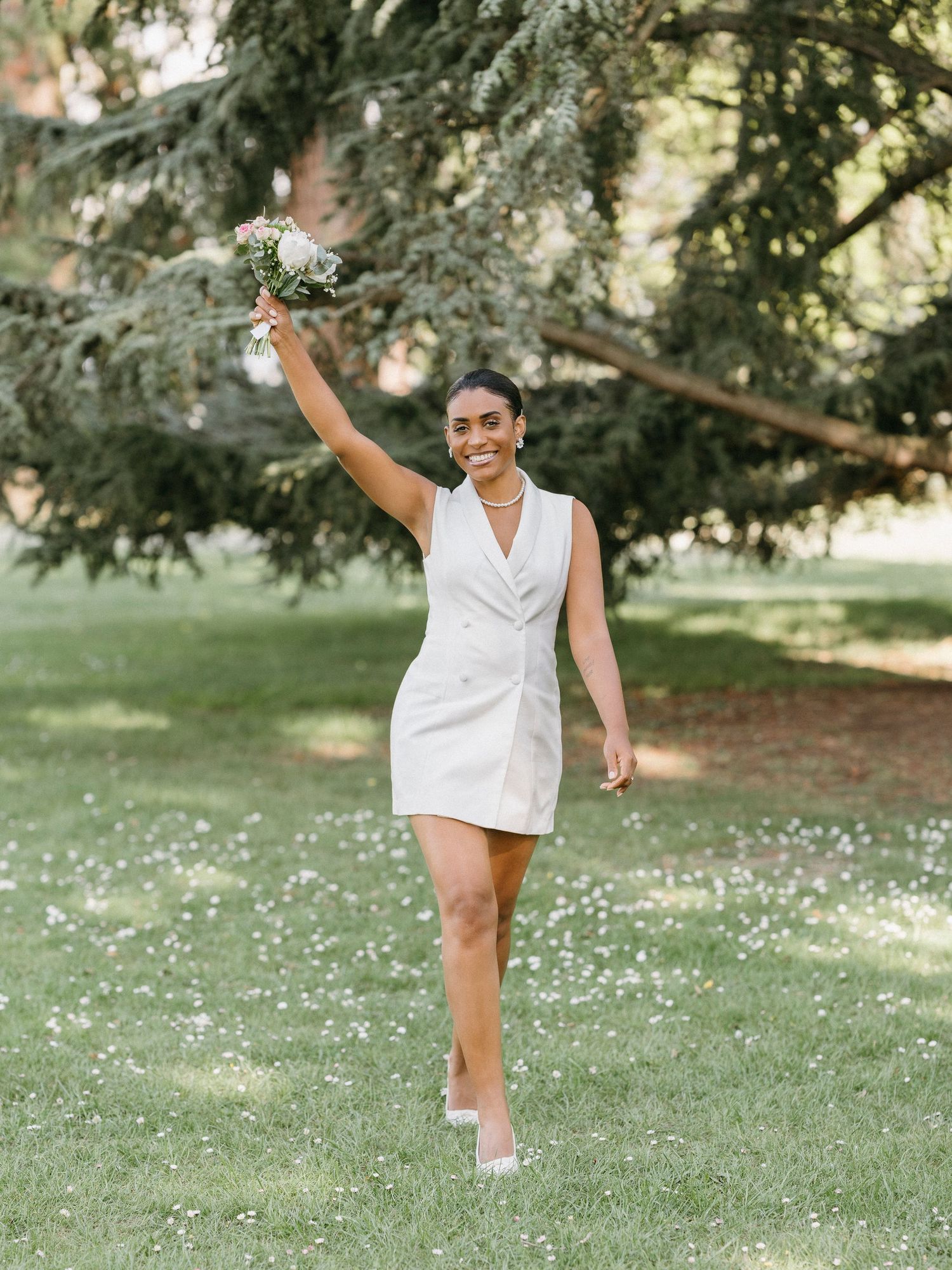 Joyful bride in white mini blazer dress raising bouquet triumphantly while walking through flower-dotted park.