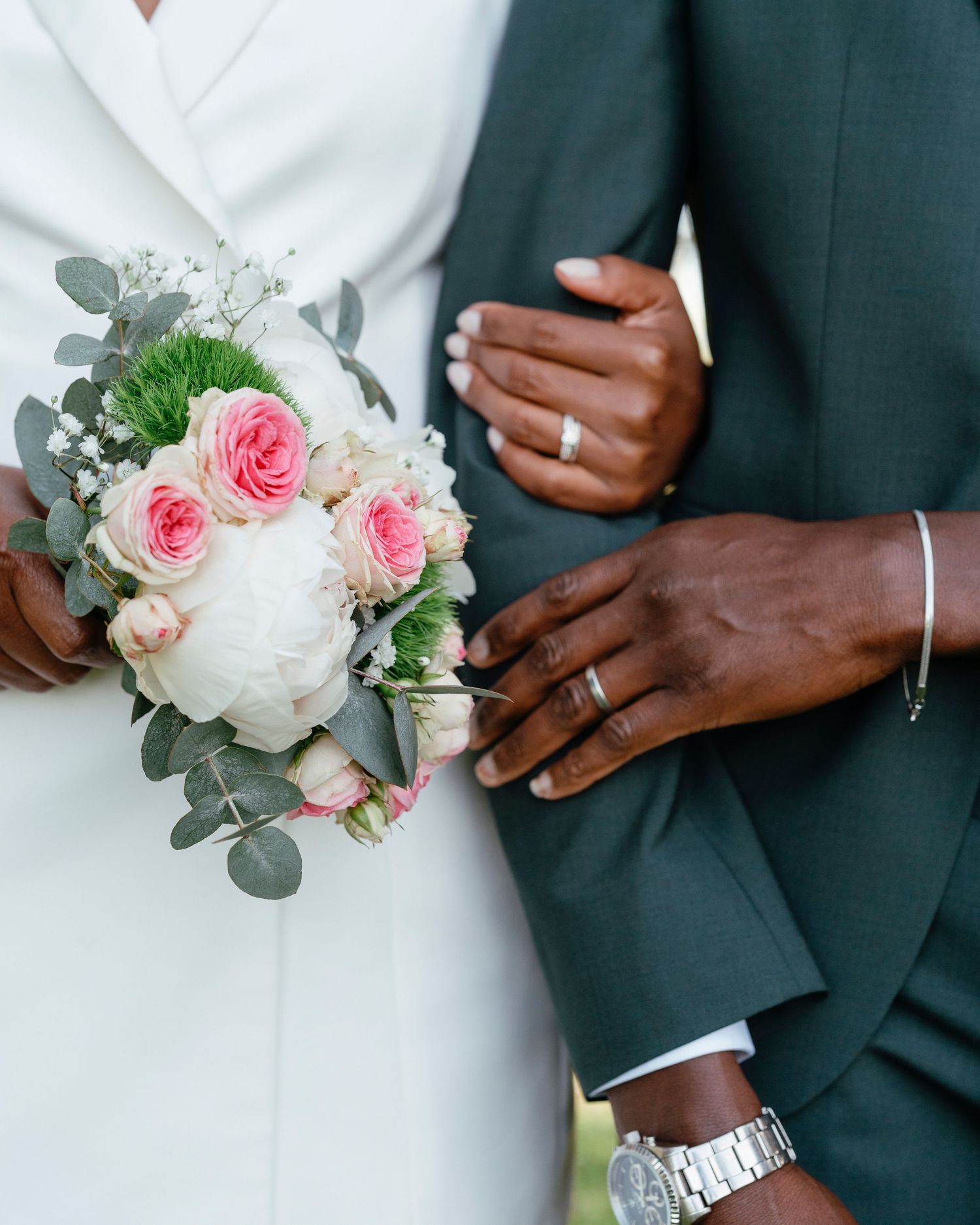 Close-up of newlyweds holding hands showing wedding rings, with bride holding a pink and white rose bouquet.