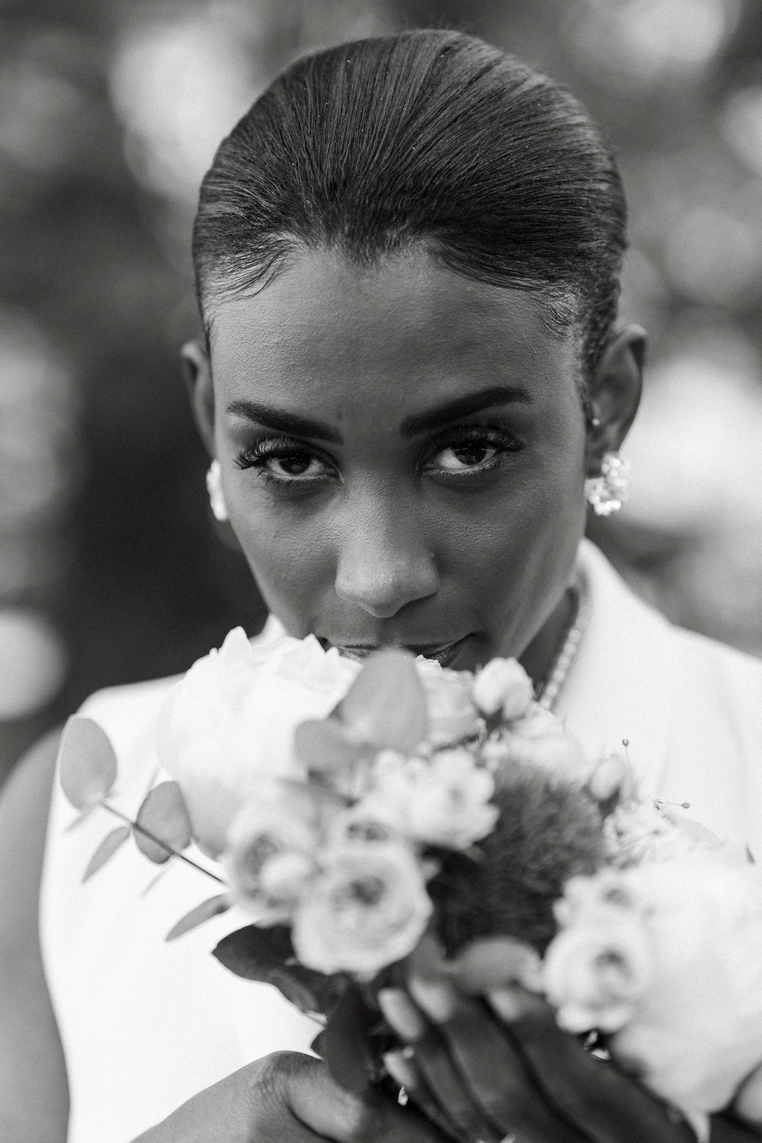 Black and white portrait of bride smelling wedding bouquet of roses and eucalyptus, wearing pearl jewelry.