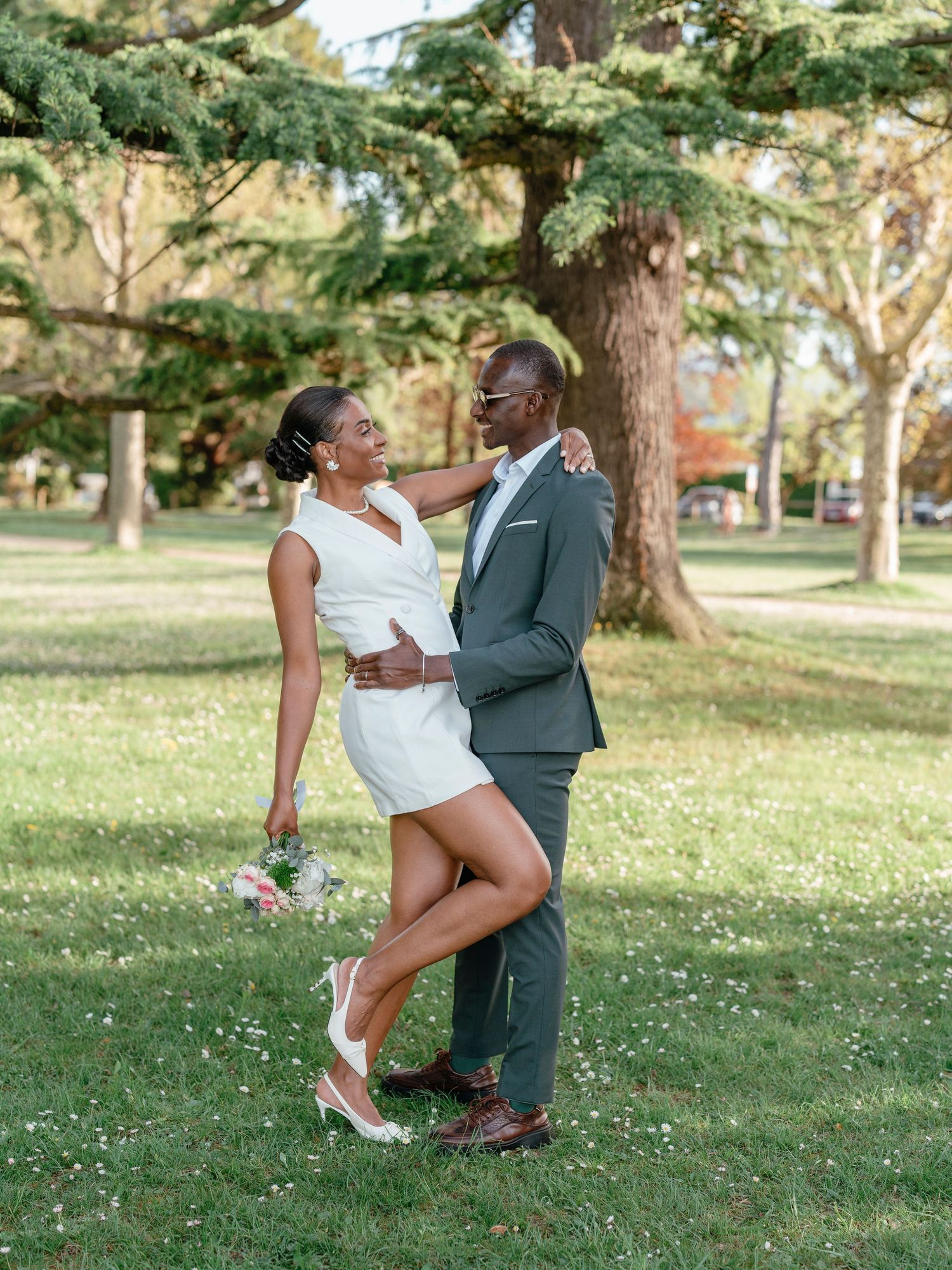 Bride in white mini dress playfully lifting her leg while embracing groom in gray suit in a green park.