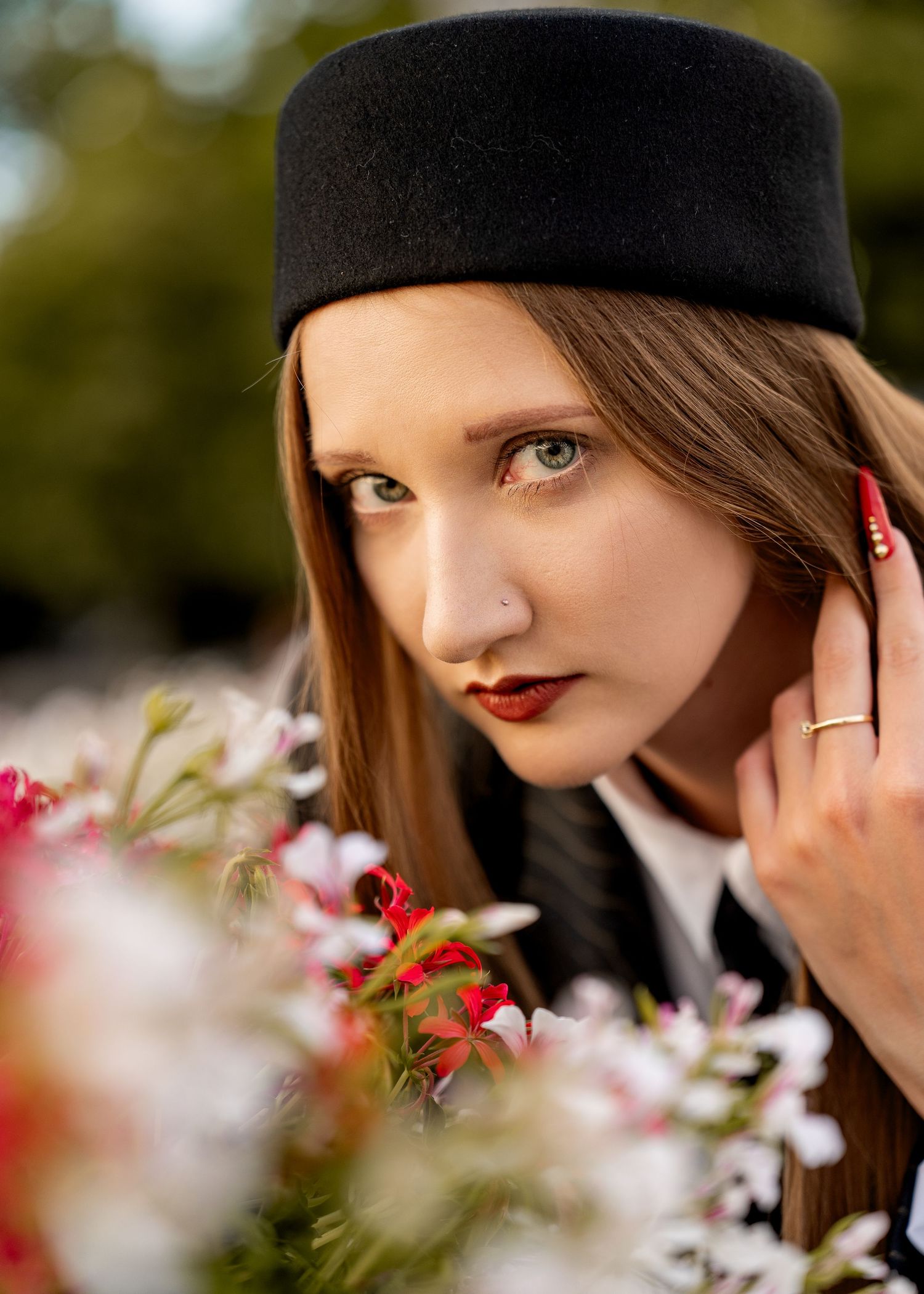 Young woman in black pillbox hat with red lips poses near colorful flowers in a elegant outdoor portrait.