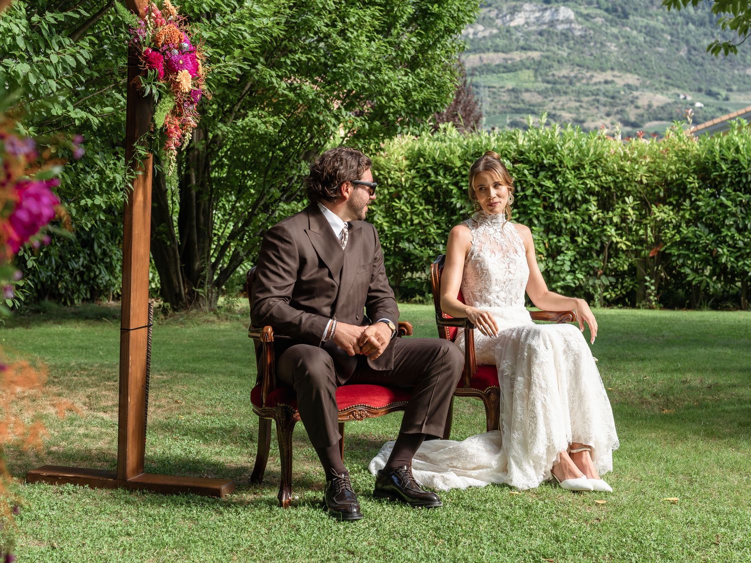 Bride and groom seated on red chairs during an outdoor wedding ceremony surrounded by lush green garden.
