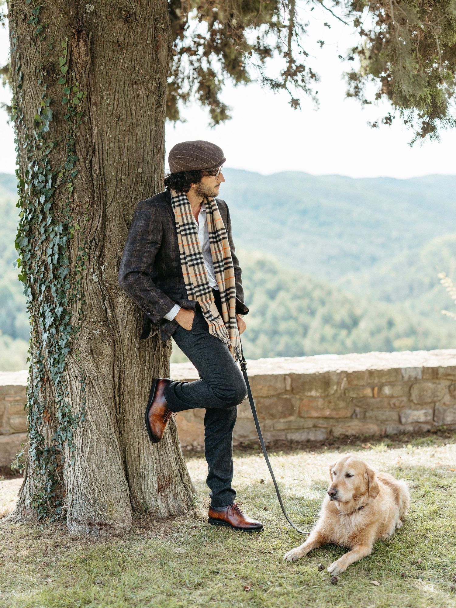 Fashionable man in plaid blazer and scarf leans on tree holding dog leash with golden retriever nearby.