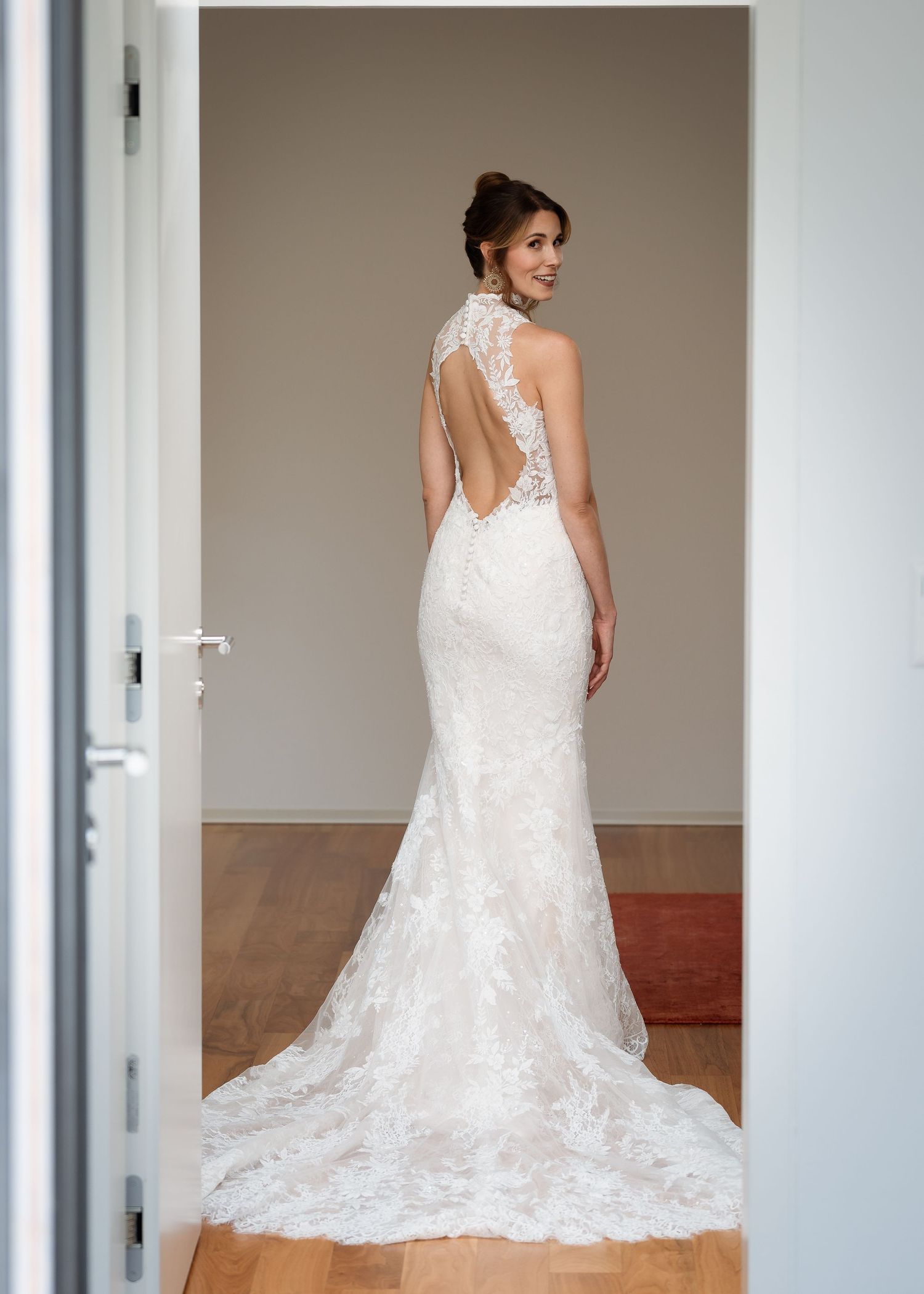 Elegant backless lace wedding gown with halter neck and train, modeled from behind in a bright doorway.