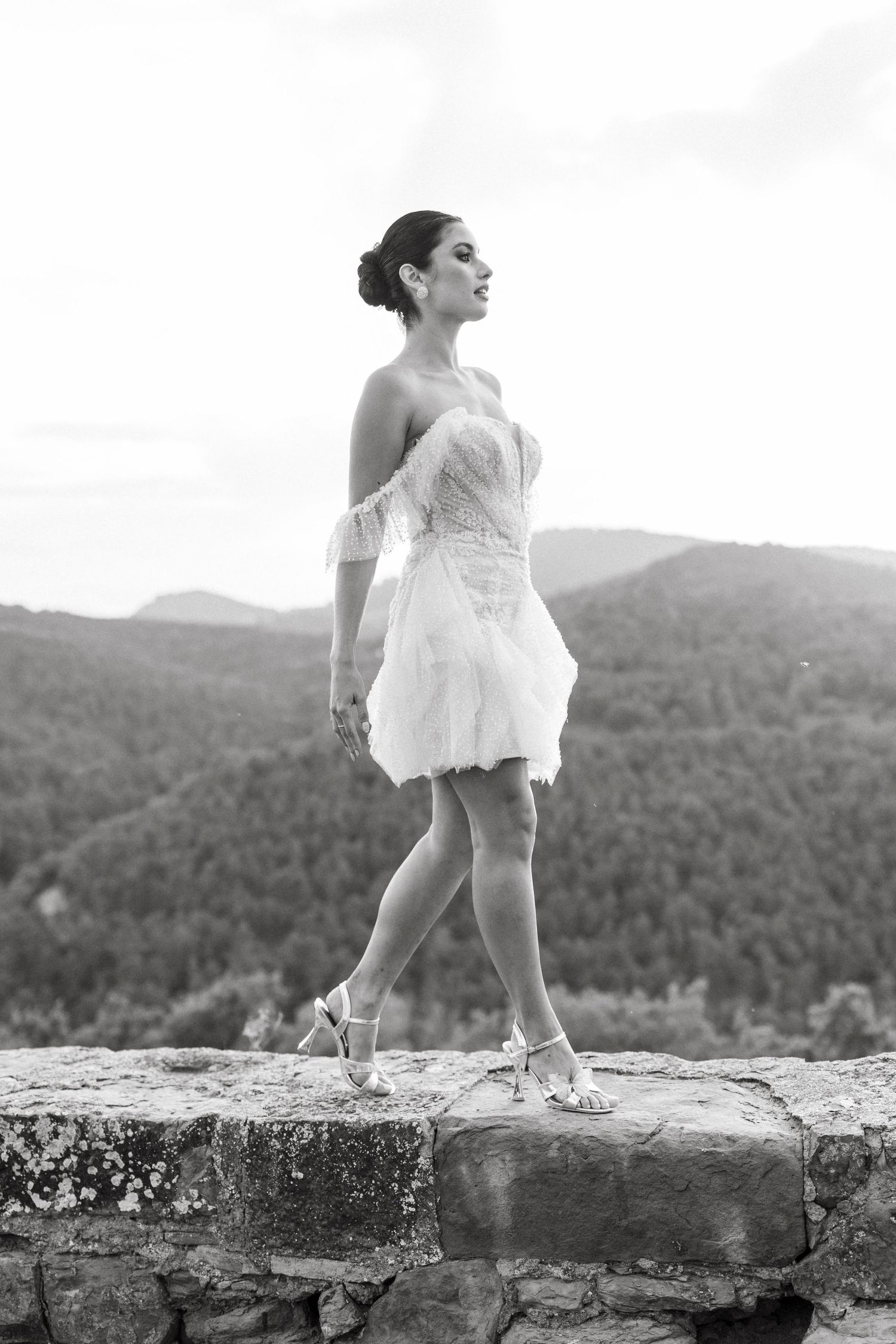 Black and white photo of a bride in a short white dress standing on a stone wall with mountain views.