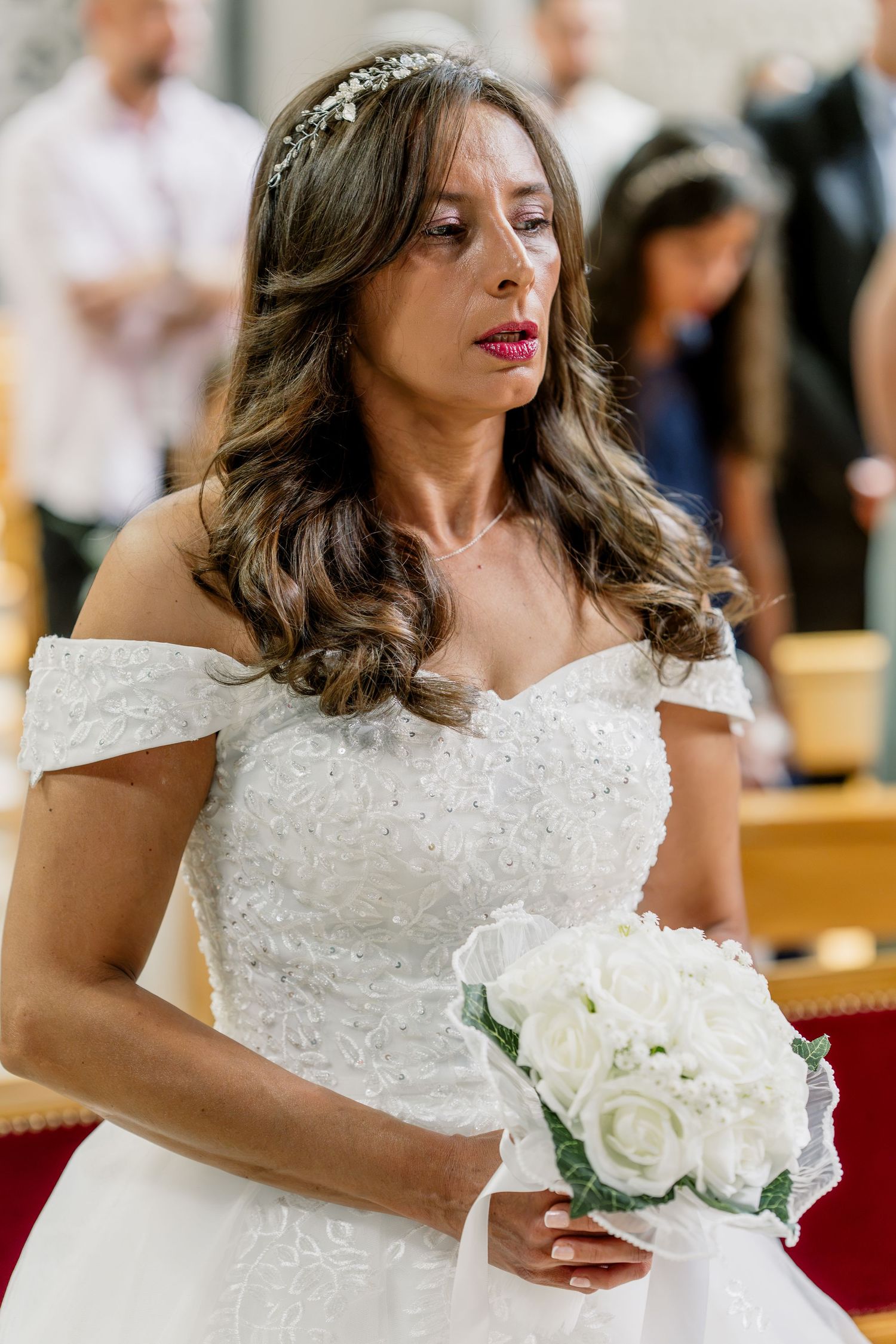 Bride in white off-shoulder lace gown holding white rose bouquet with floral hair accessory at church ceremony.