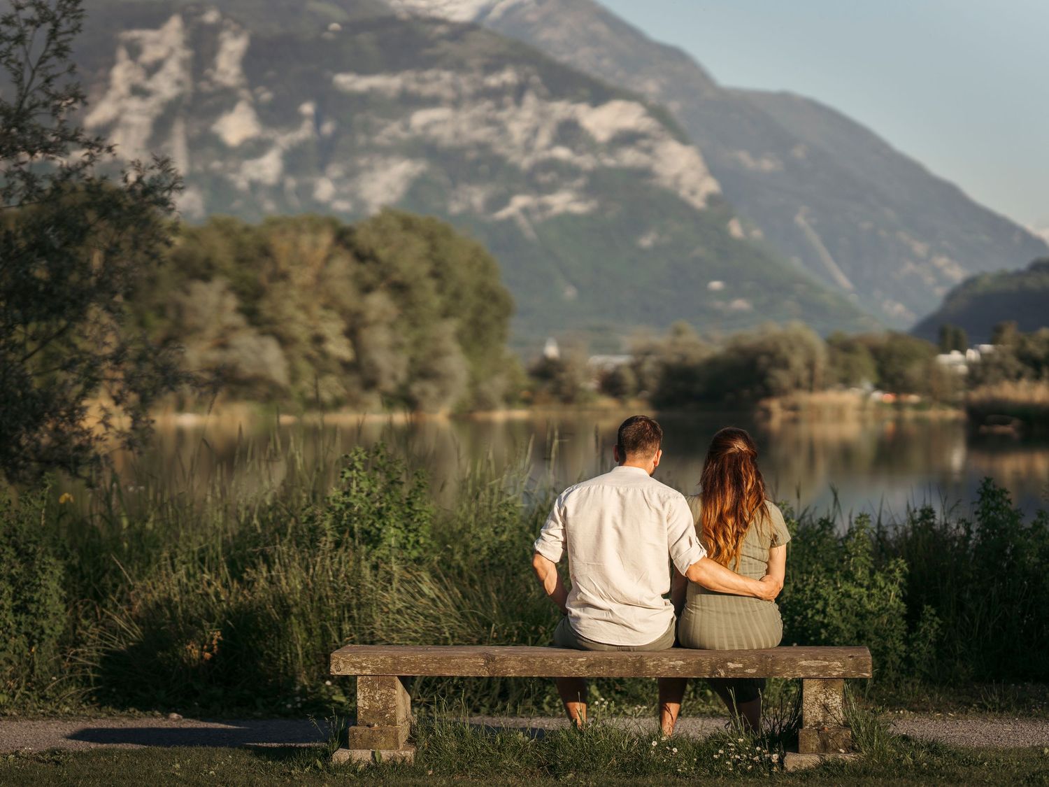 Couple sitting together on a wooden bench by a scenic mountain lake surrounded by lush greenery.
