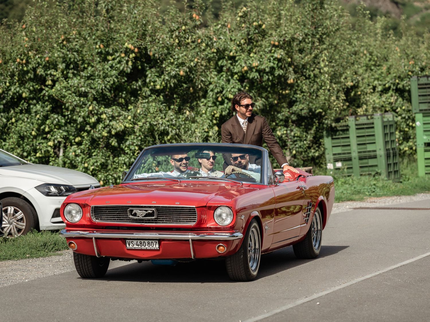 A man in a suit drives a classic red Ford Mustang convertible on a sunny day.