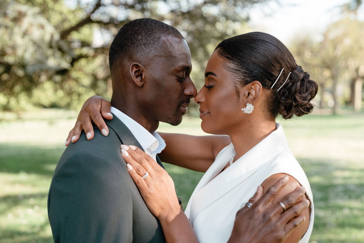 A couple in wedding attire share a tender nose-to-nose moment outdoors in a sunlit park setting.