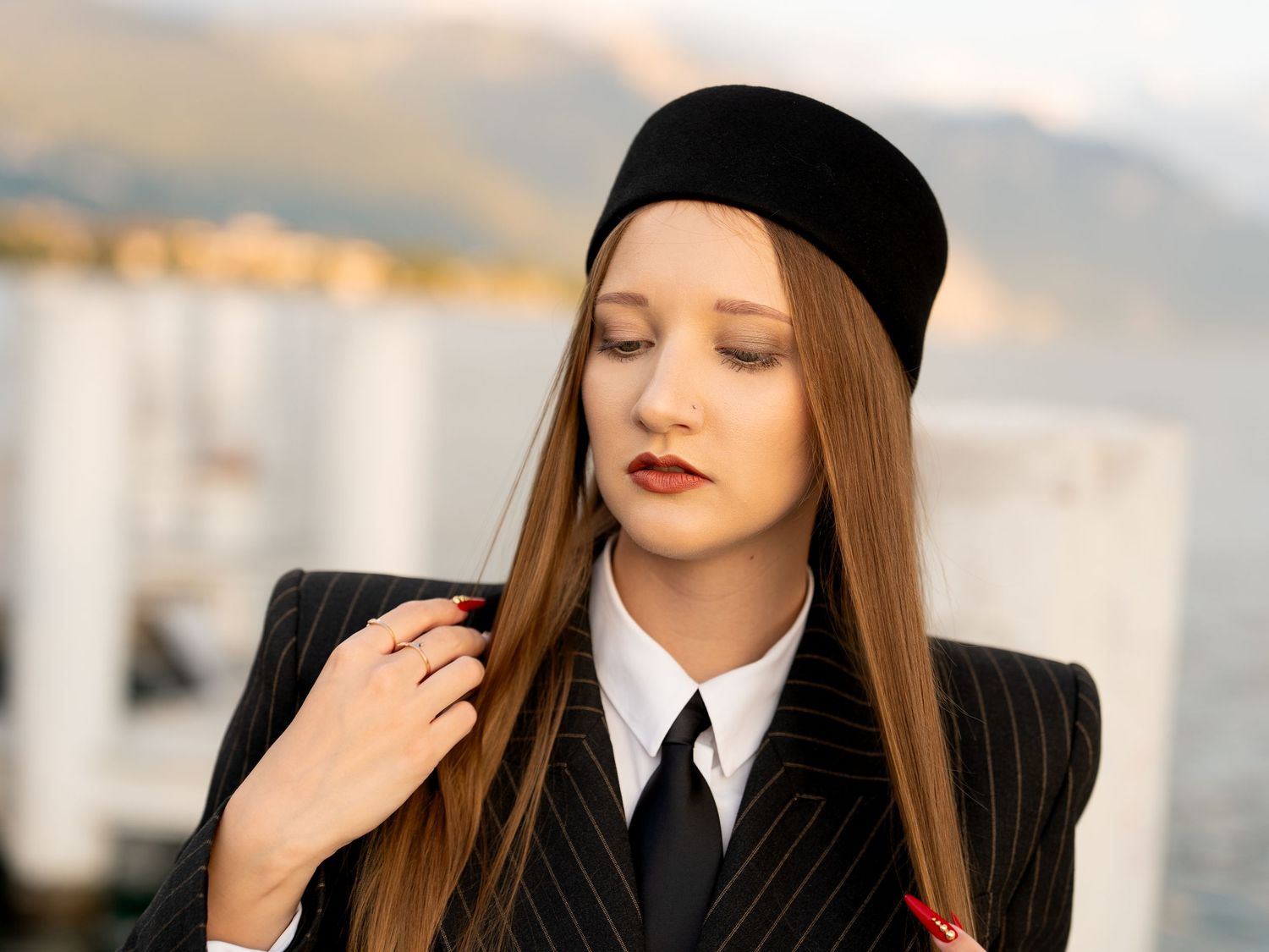 Young woman in black pinstripe blazer, tie, white shirt, and beret hat poses outdoors near waterfront.