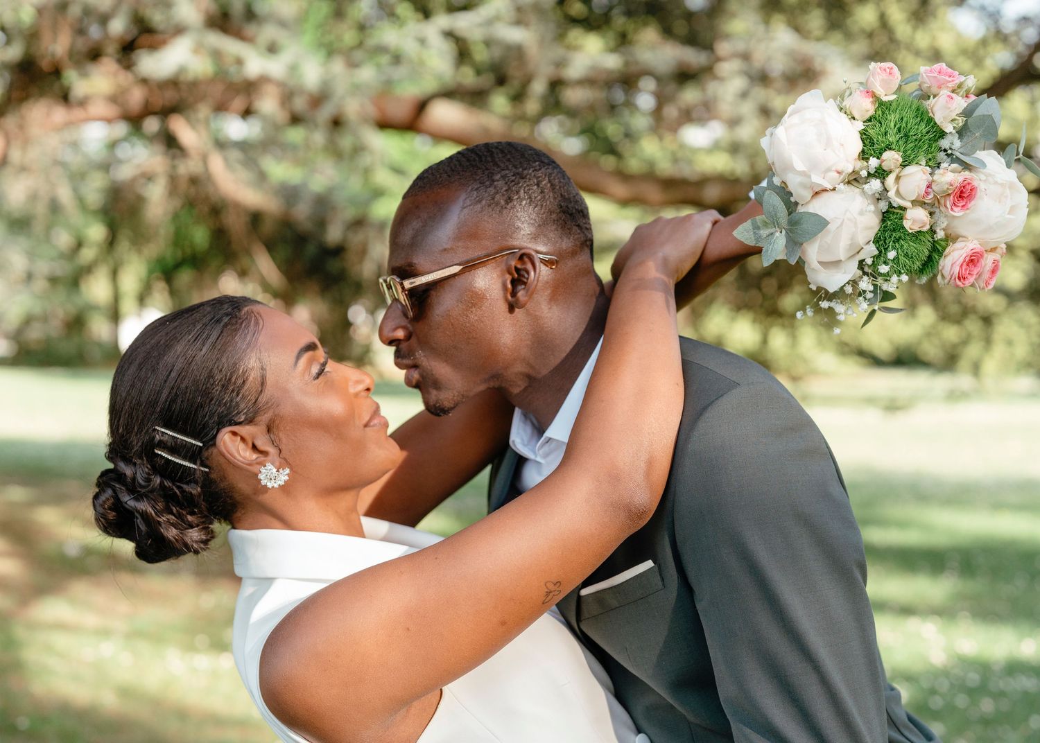 Bride and groom sharing a kiss outdoors, groom in gray suit holding a pink and white rose wedding bouquet.