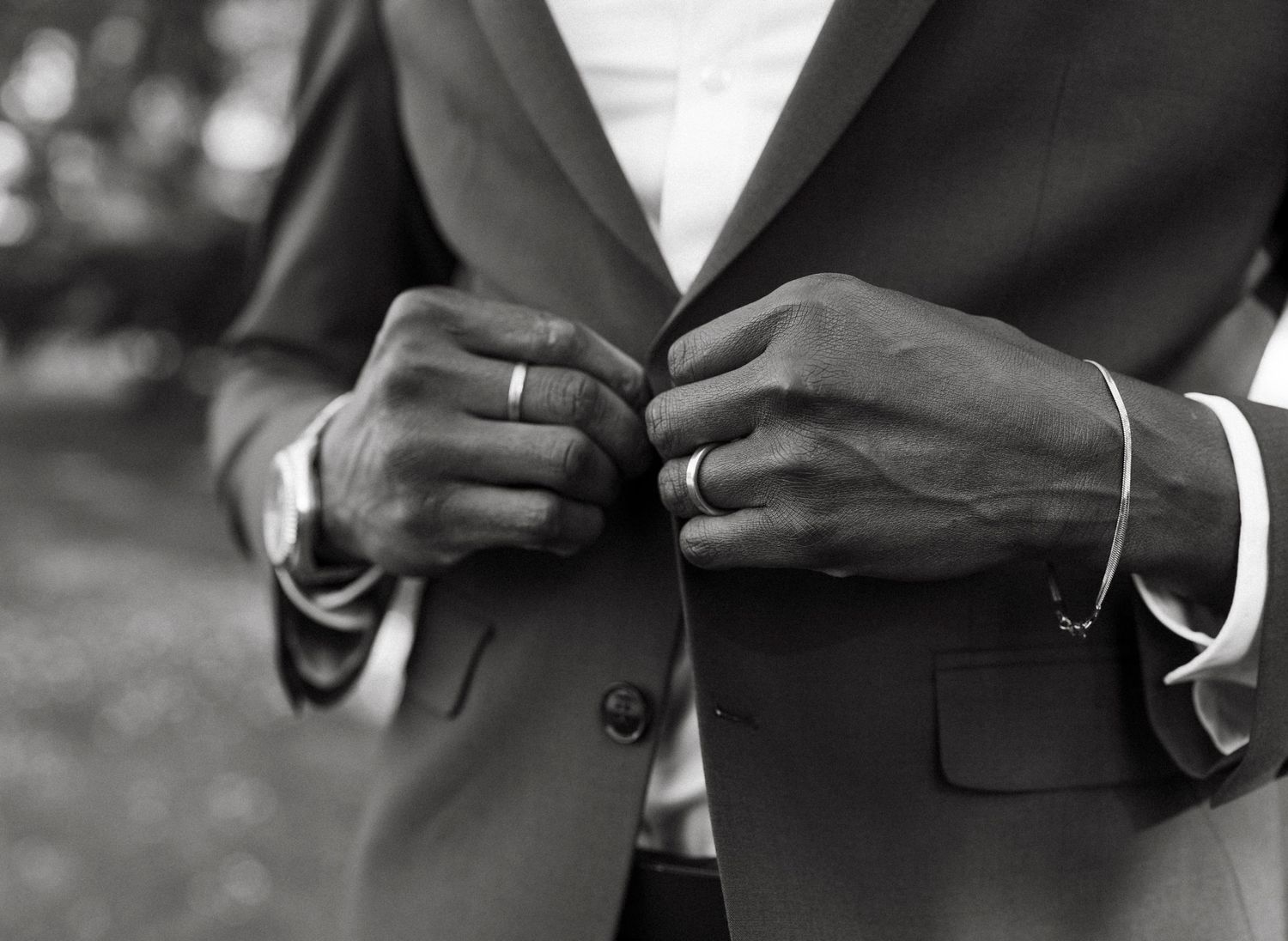 Close-up of groom's hands buttoning a dark suit jacket with wedding rings visible.
