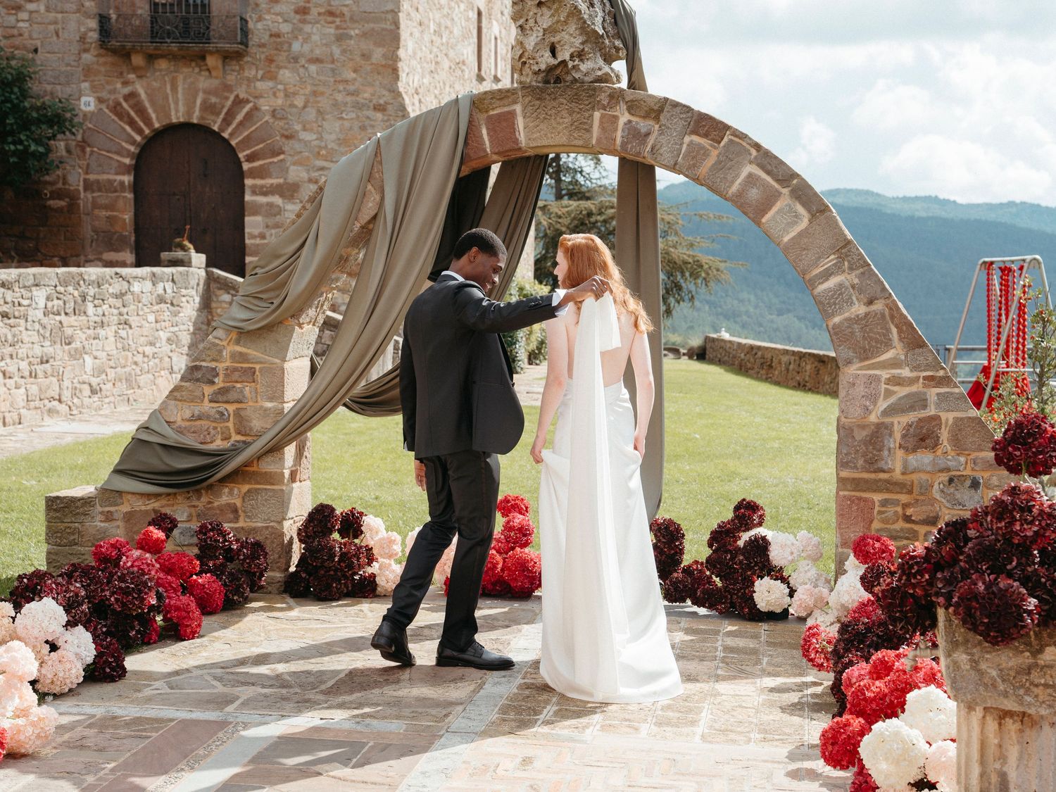 Couple exchanging vows under a stone arch adorned with red and pink flowers at an Italian countryside wedding.