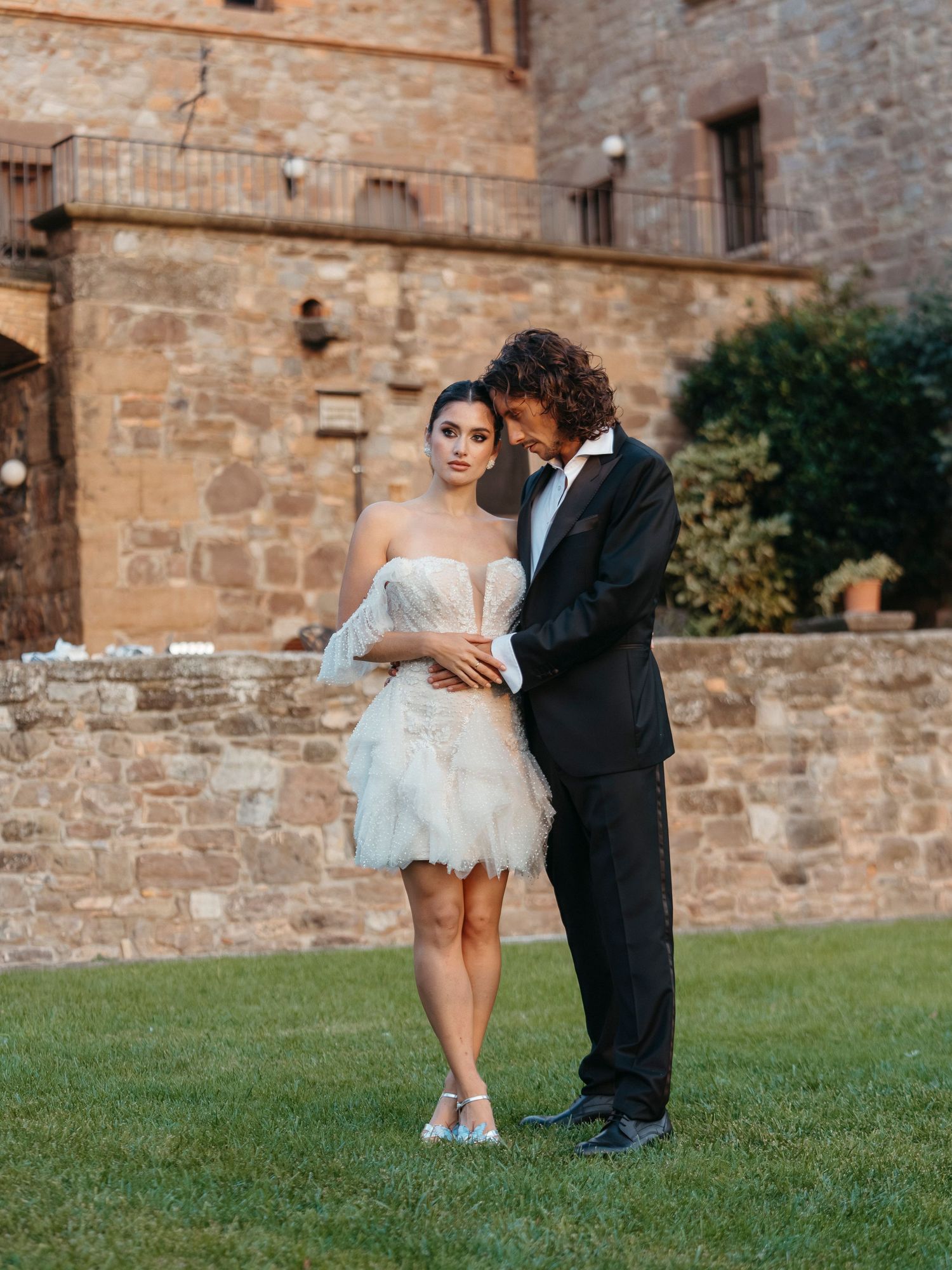 Couple in wedding attire pose before stone castle, bride in short feathered gown, groom in black tuxedo.