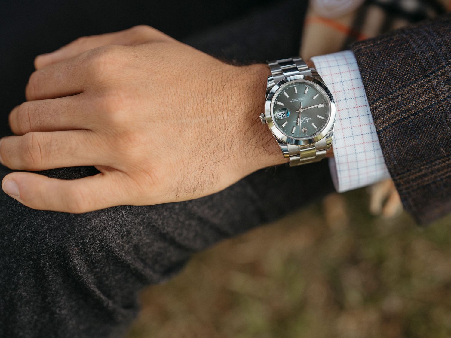 Close-up of a mans wrist wearing a silver Rolex watch with a gray dial, paired with a plaid suit jacket.