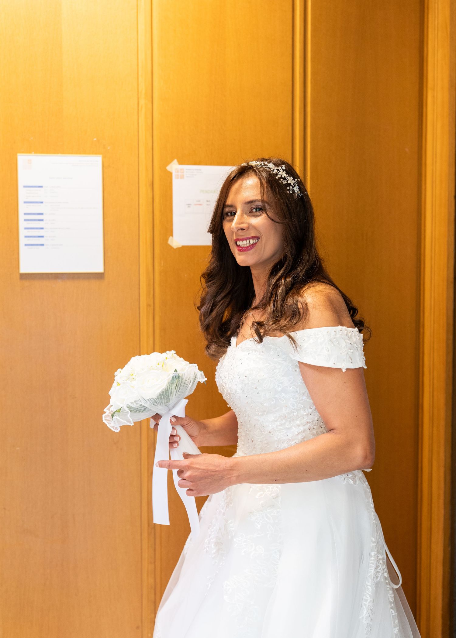 Smiling bride in white off-shoulder gown holding bouquet of white roses, wearing pearl hair accessory.