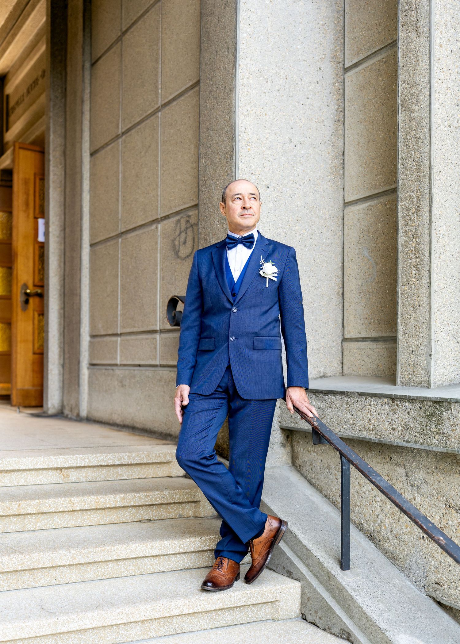 Groom in navy blue suit with bow tie and boutonniere posing on stone steps outside a classic building.