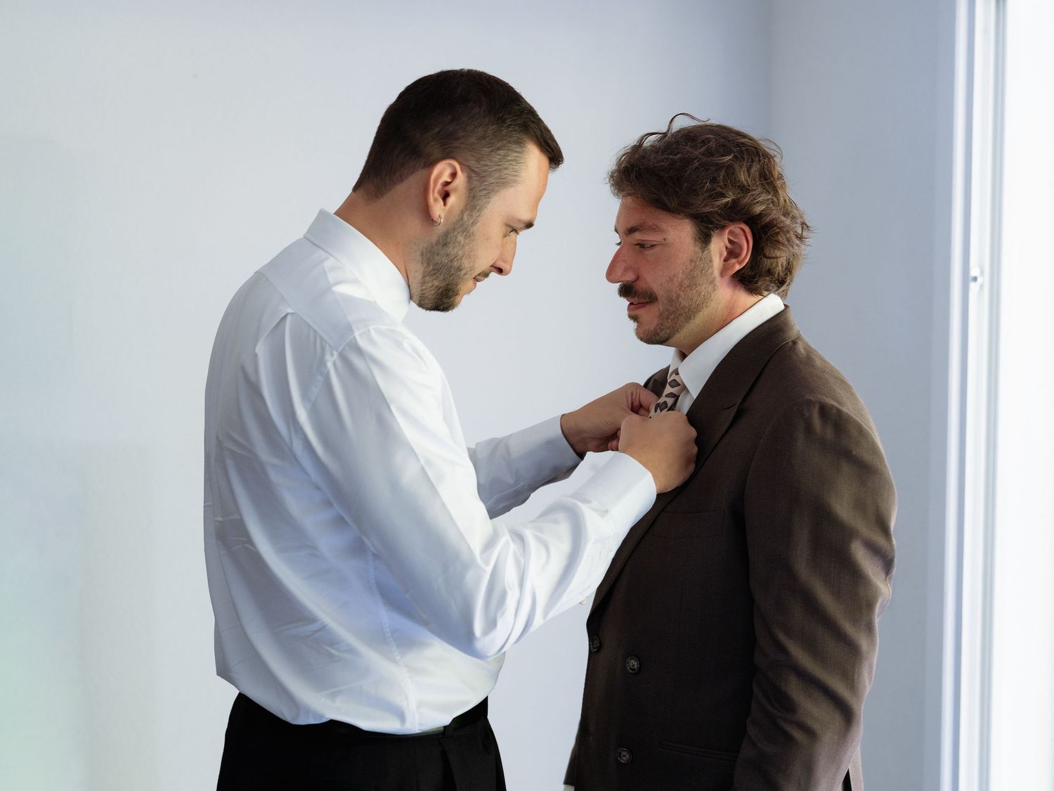Man in white shirt adjusting tie of another man in brown suit jacket against white background.