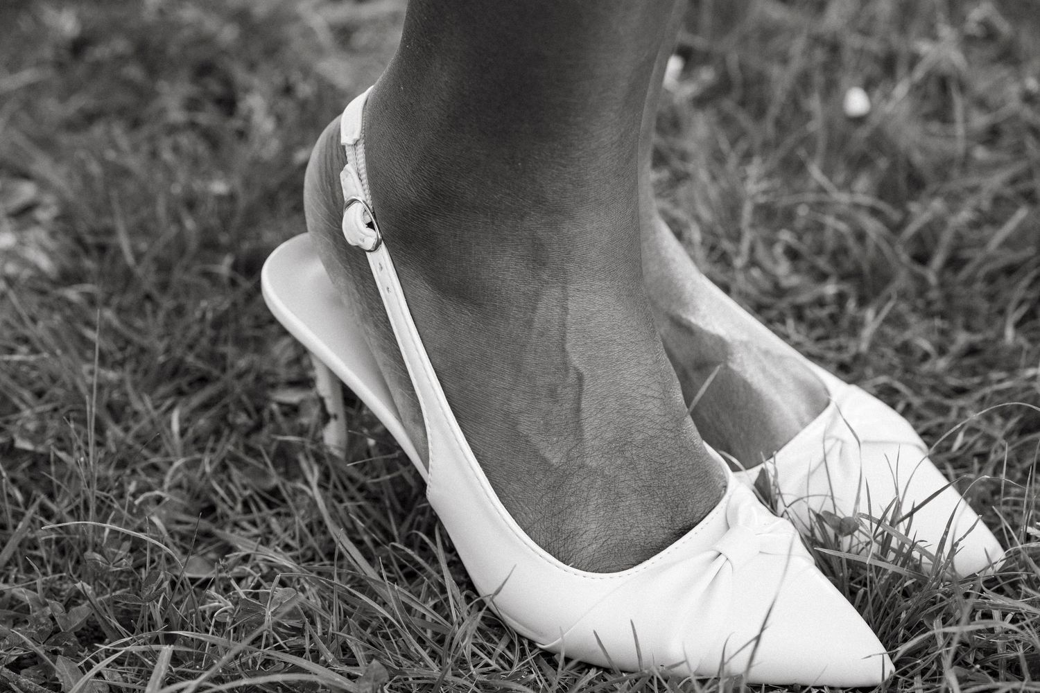 Black and white close-up of bride wearing white pointed-toe slingback heels on grass.