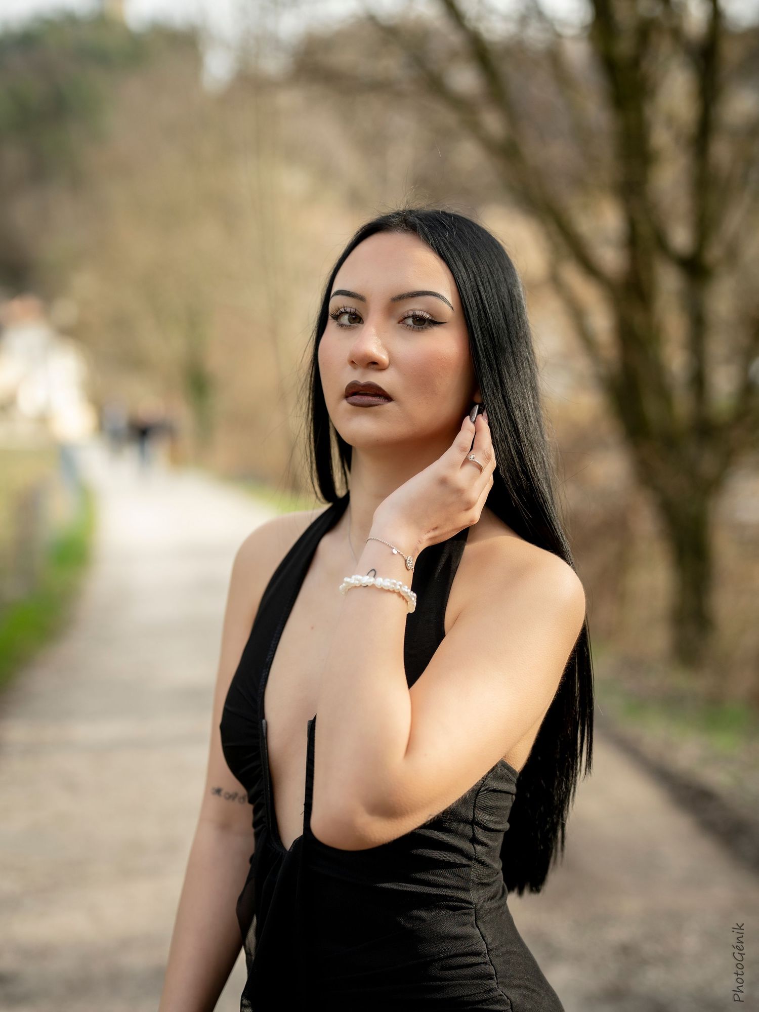 Portrait of a woman with long straight dark hair in a black dress touching her neck outdoors.