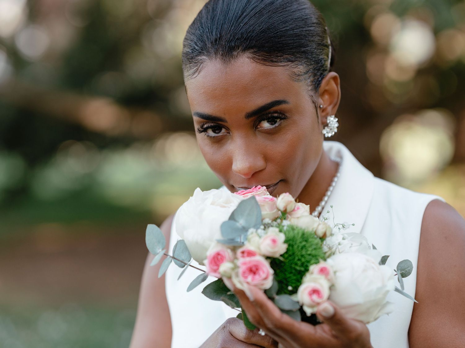 Bride in white blazer dress holding bouquet close to face, smelling fresh pink and white roses.