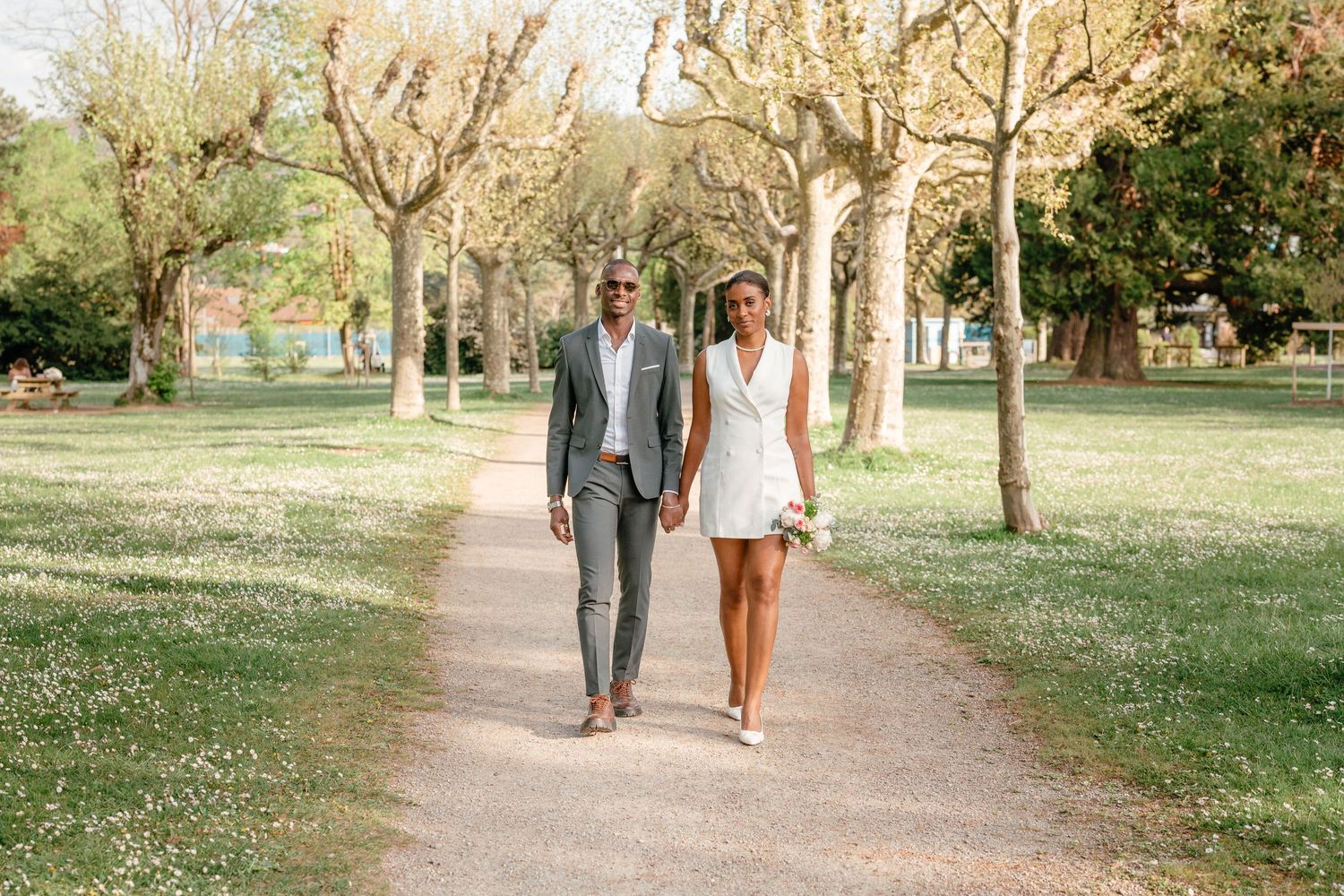 Elegant couple walking hand in hand along a tree-lined park path on their wedding day.