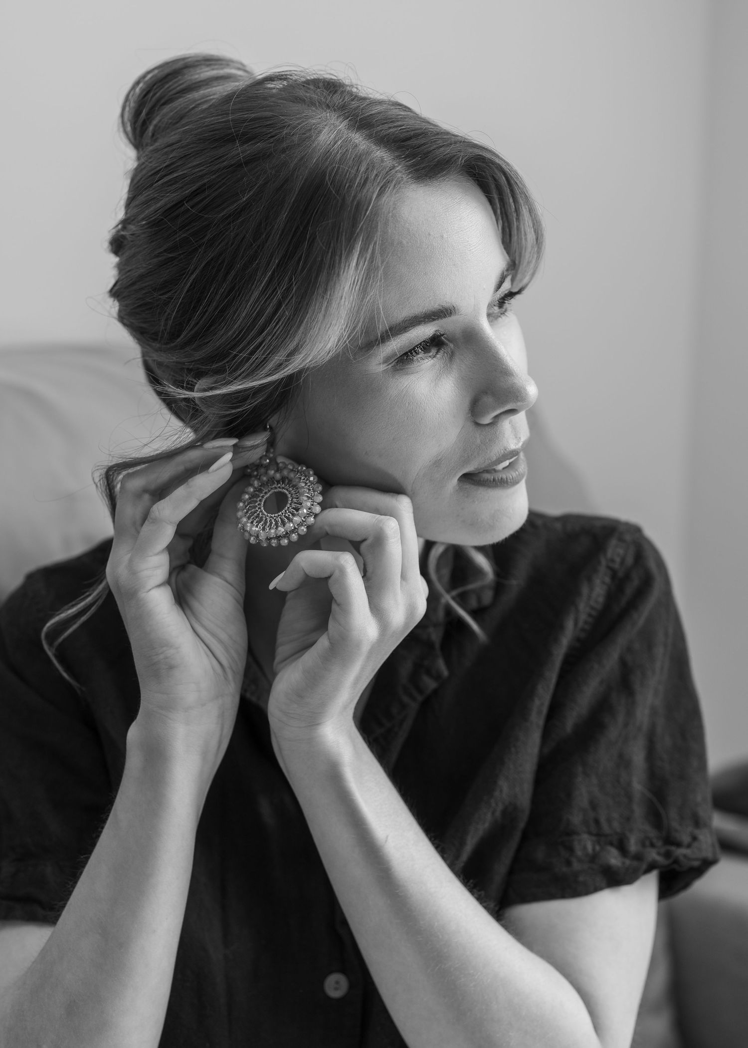 Black and white portrait of a woman touching her decorative round earring, hair up, wearing a dark top.