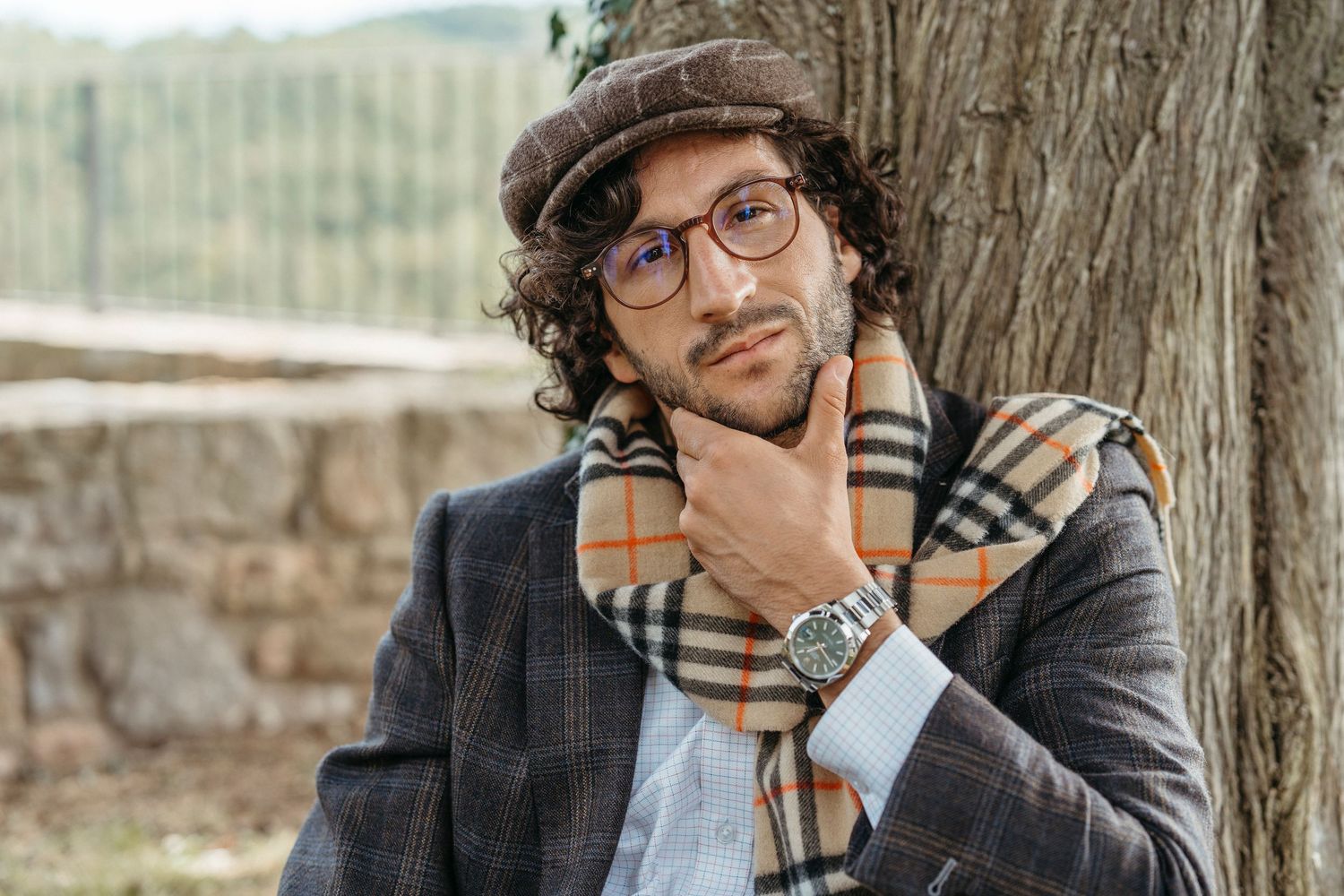 Stylish man in flat cap and Burberry scarf poses thoughtfully against a tree in autumn setting.
