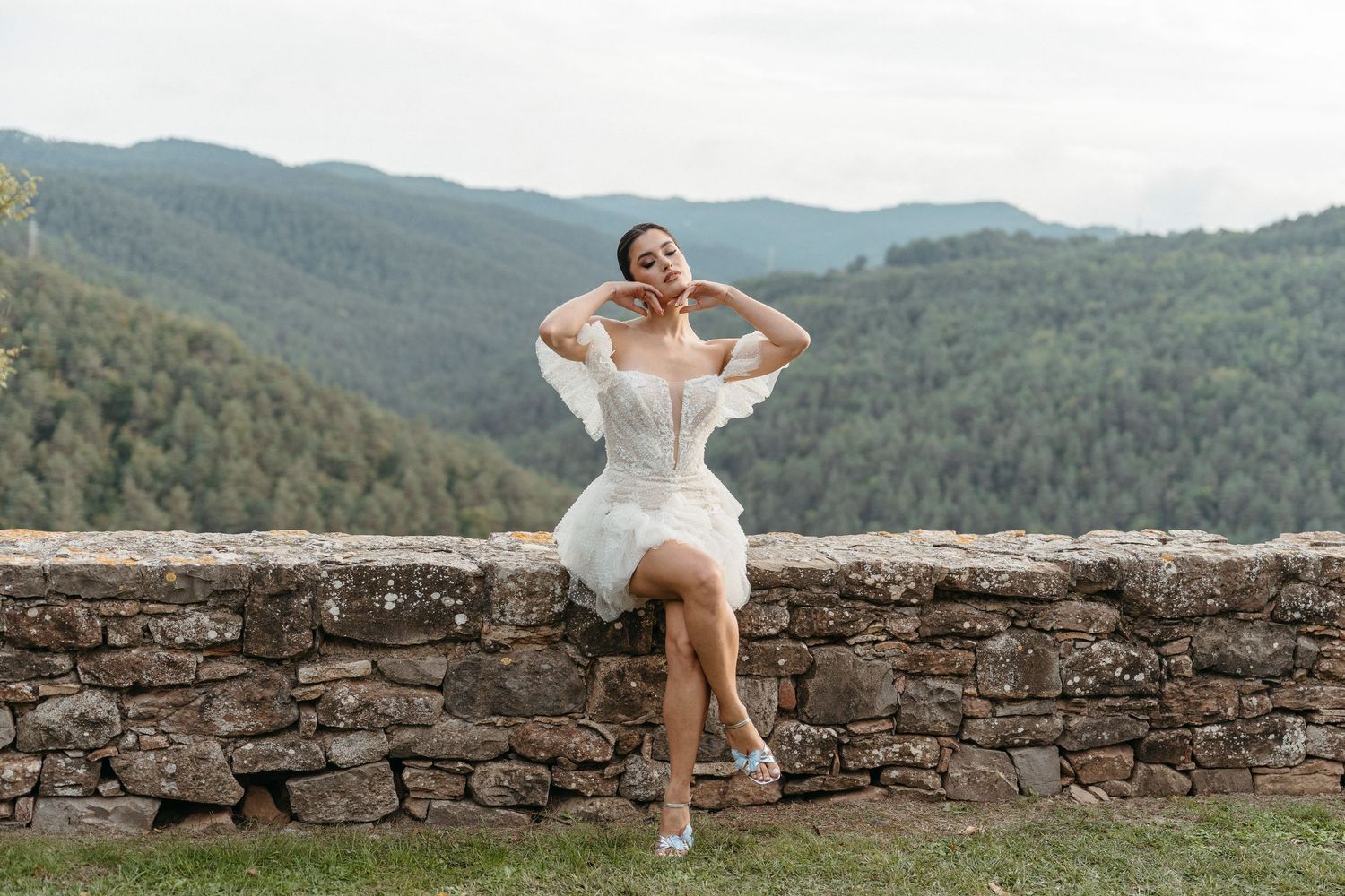 Bride in short white lace dress sits on stone wall with lush green mountain landscape backdrop.