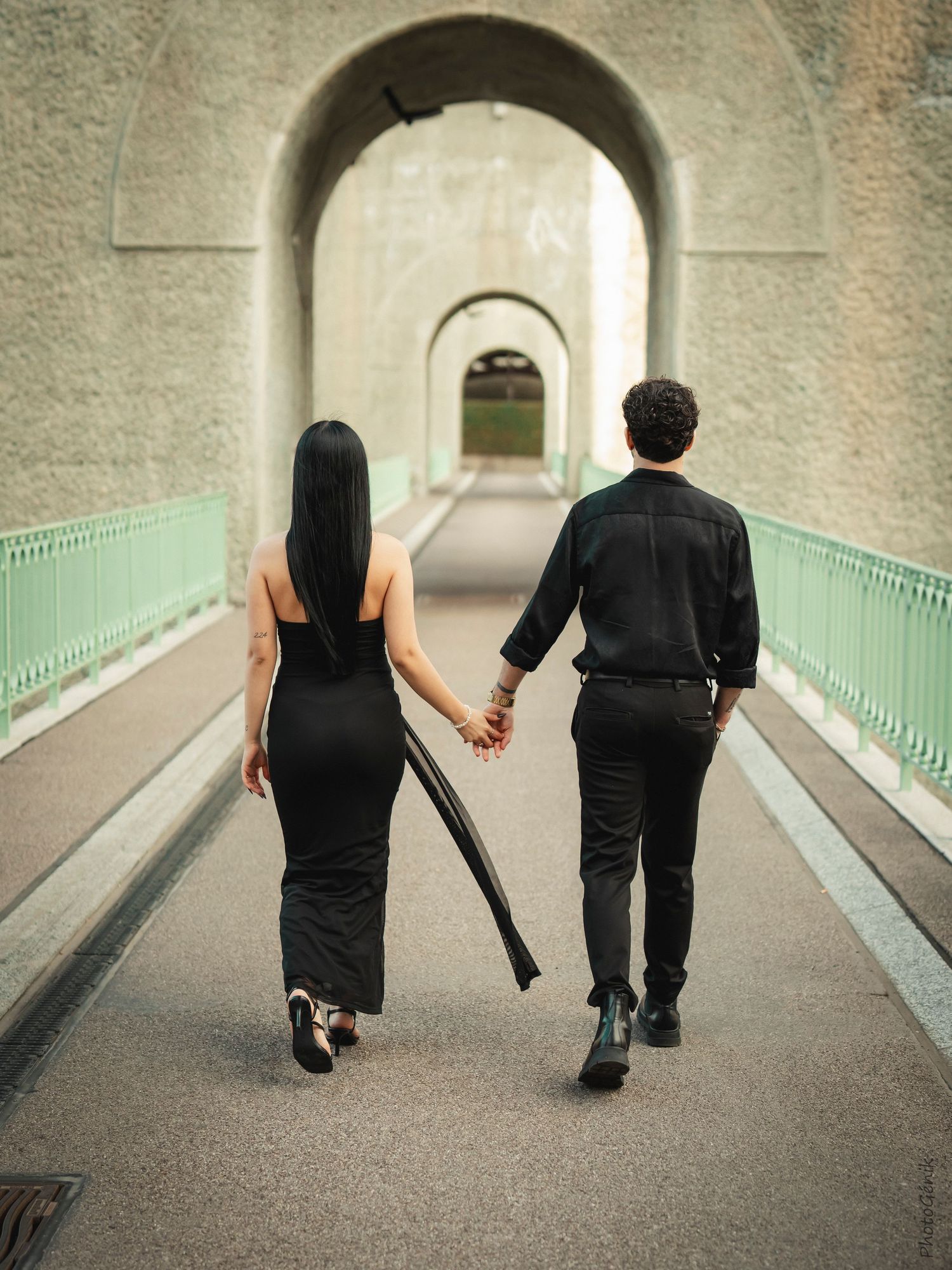 A couple dressed in black walk hand in hand through an arched tunnel bridge from behind.