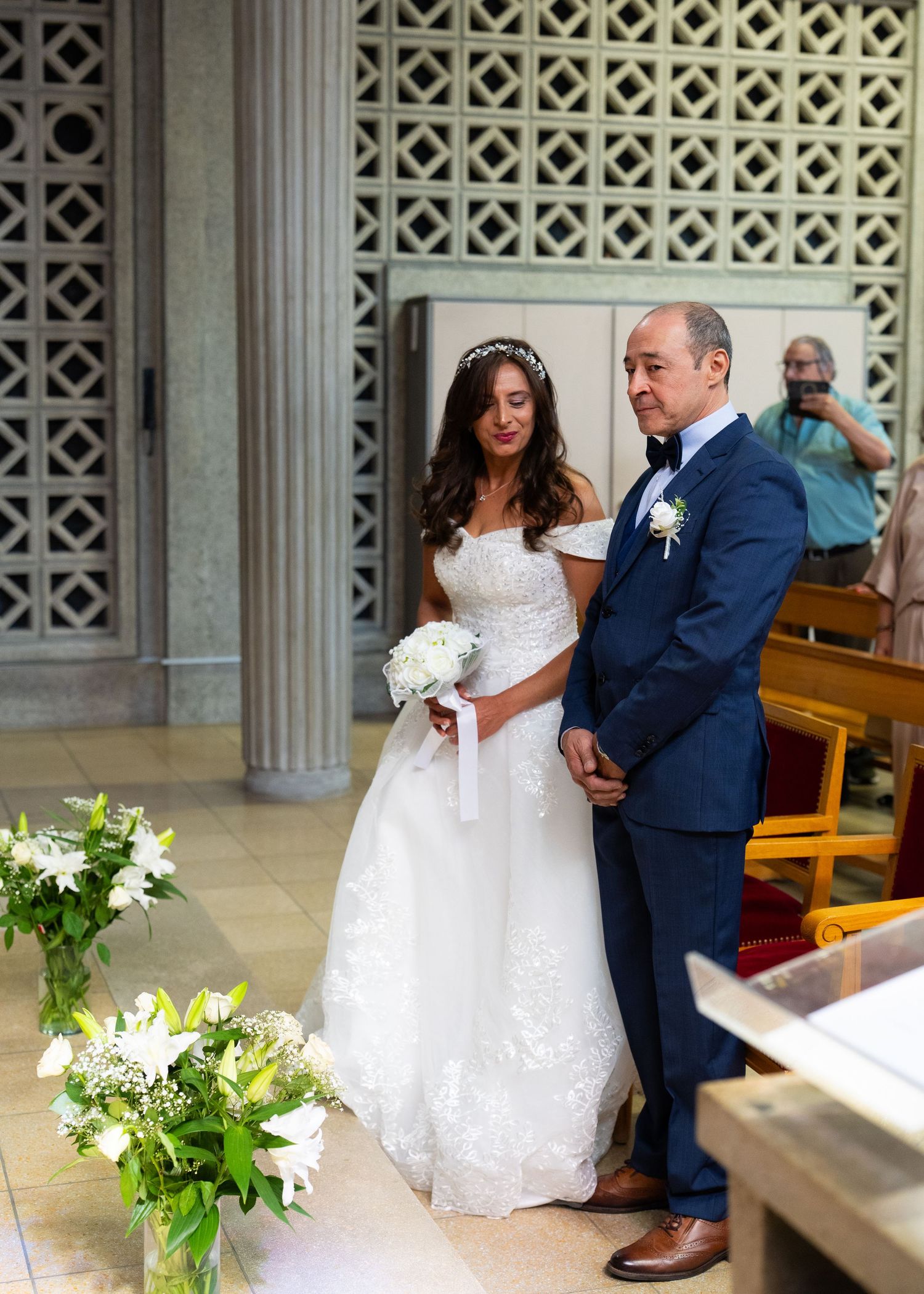 Bride in white lace gown holds bouquet beside man in blue suit during wedding ceremony in elegant church.