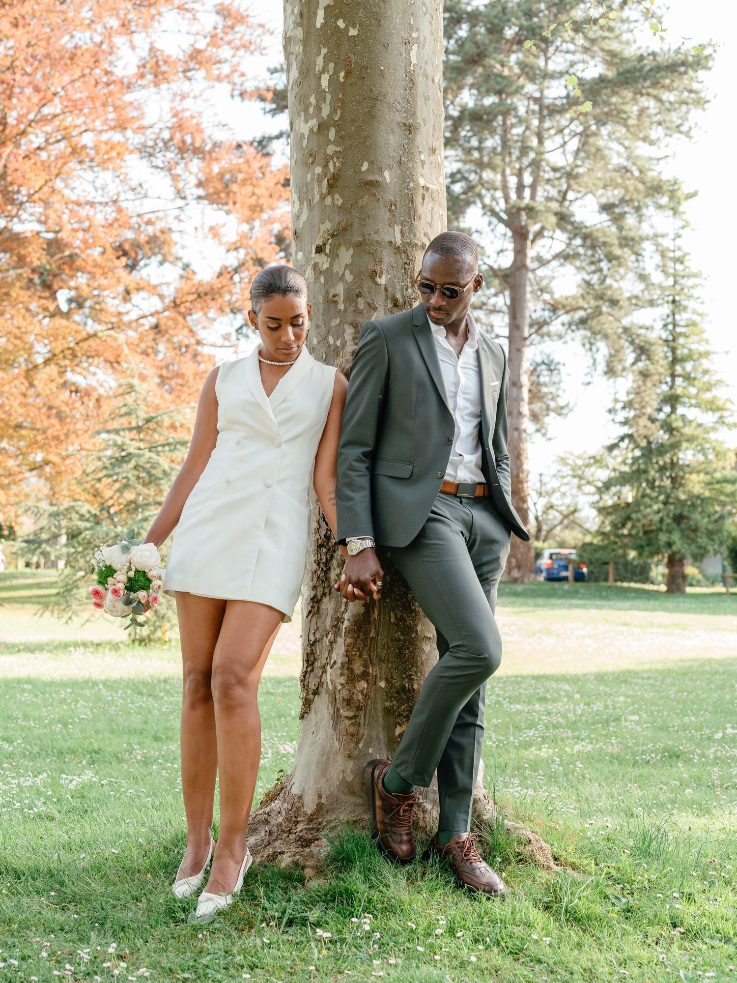 Elegant wedding couple posing by a tree in a park, bride in white mini dress, groom in grey suit.