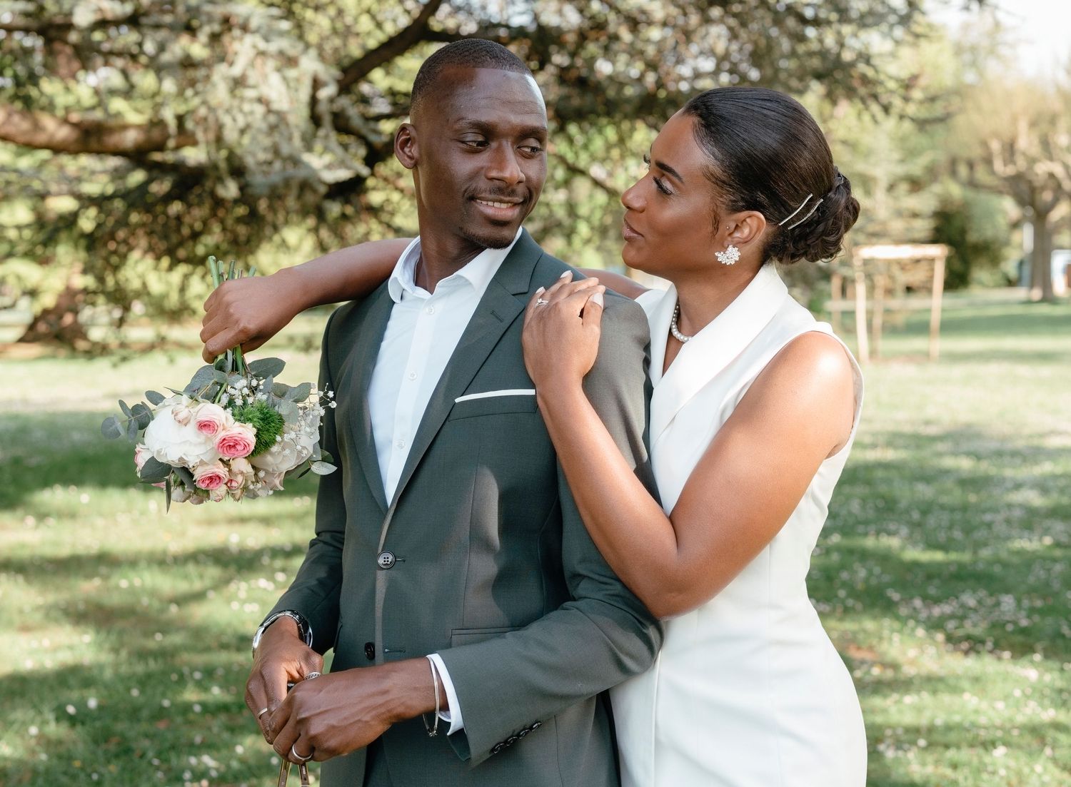 Black bride adjusting groom's suit lapel tenderly in a sunlit outdoor garden setting.