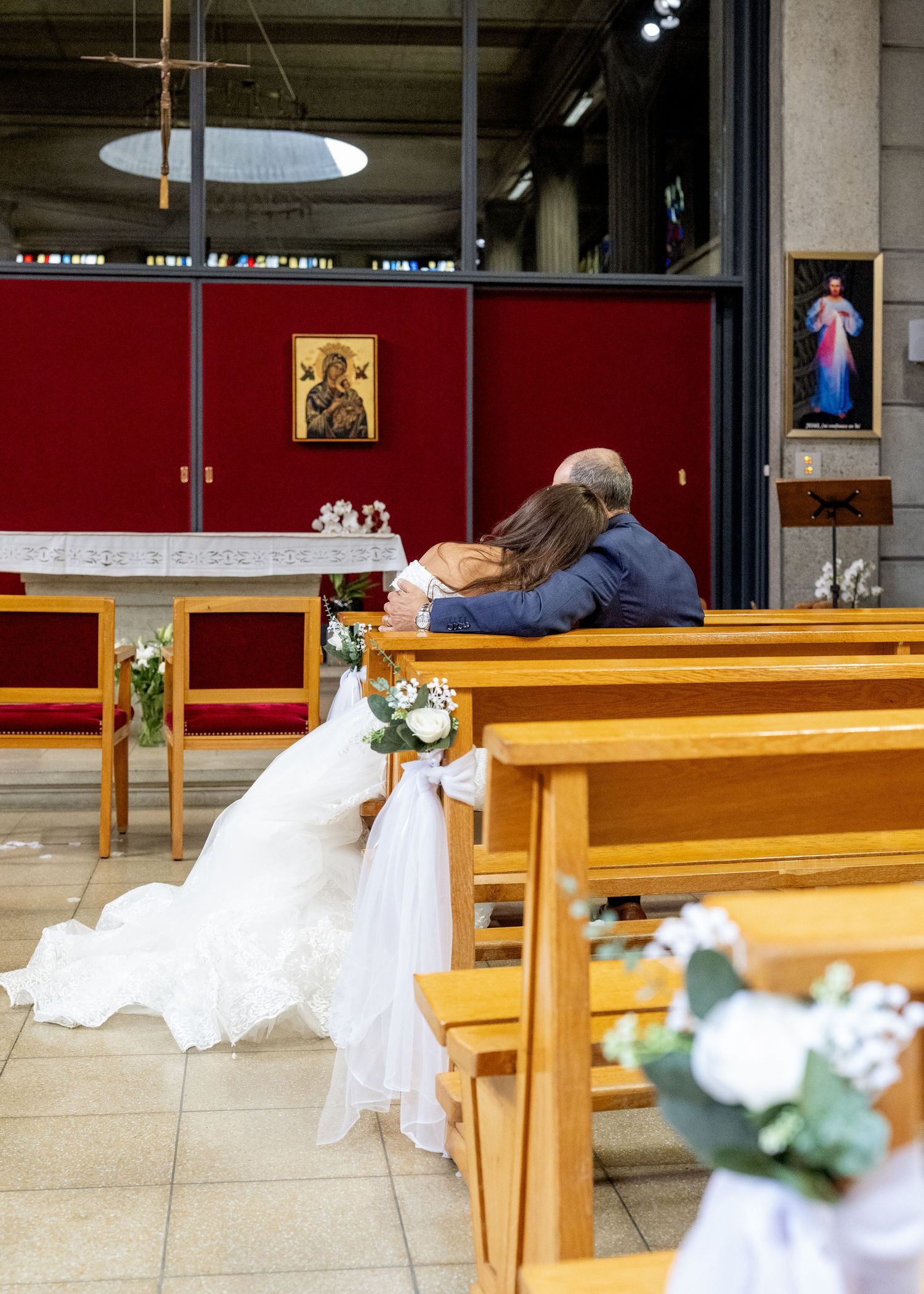 Bride and groom sitting together on a church pew during their wedding ceremony, with floral decorations and religious icons.