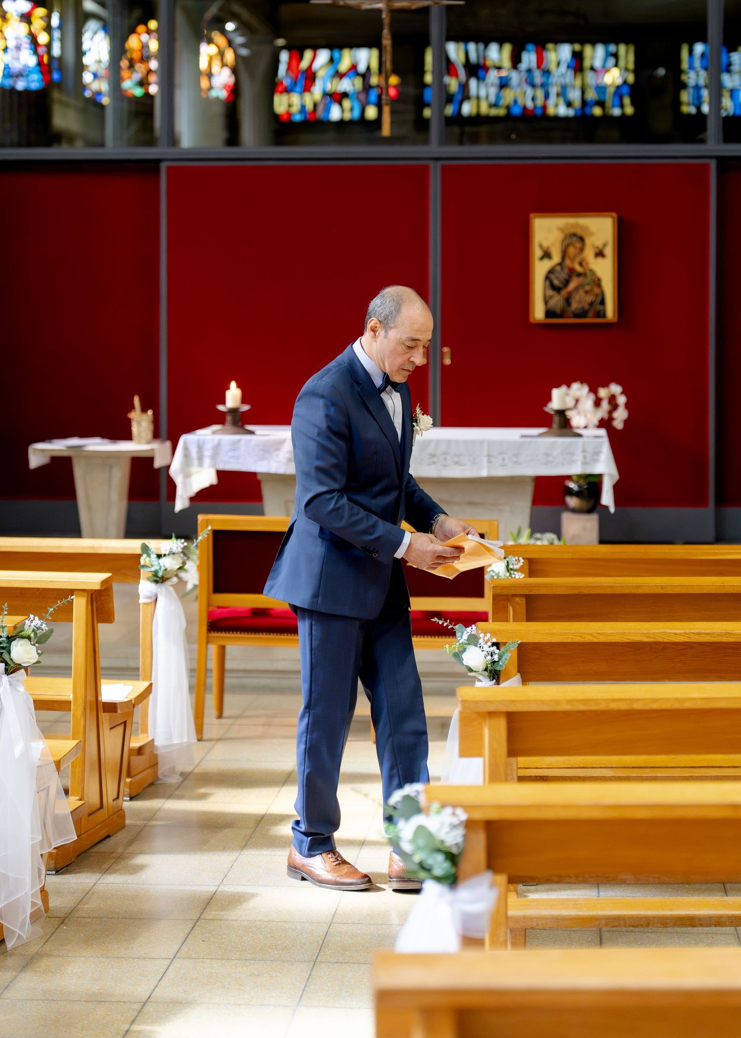 A man in a blue suit arranges items on wooden church pews decorated with white flowers before a wedding ceremony.