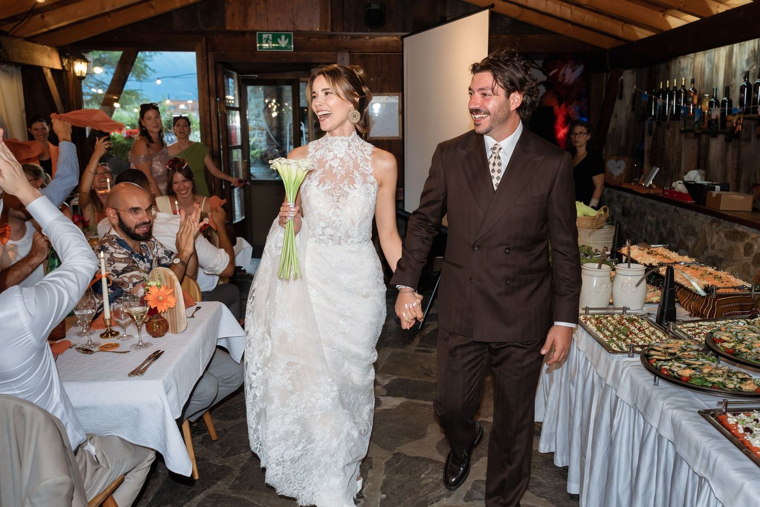 Bride in white lace gown and groom in brown suit hold hands walking into wedding reception hall.