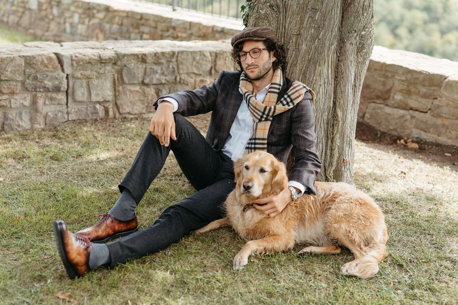 Man in plaid scarf and flat cap sits on grass with golden retriever dog beside a tree outdoors.