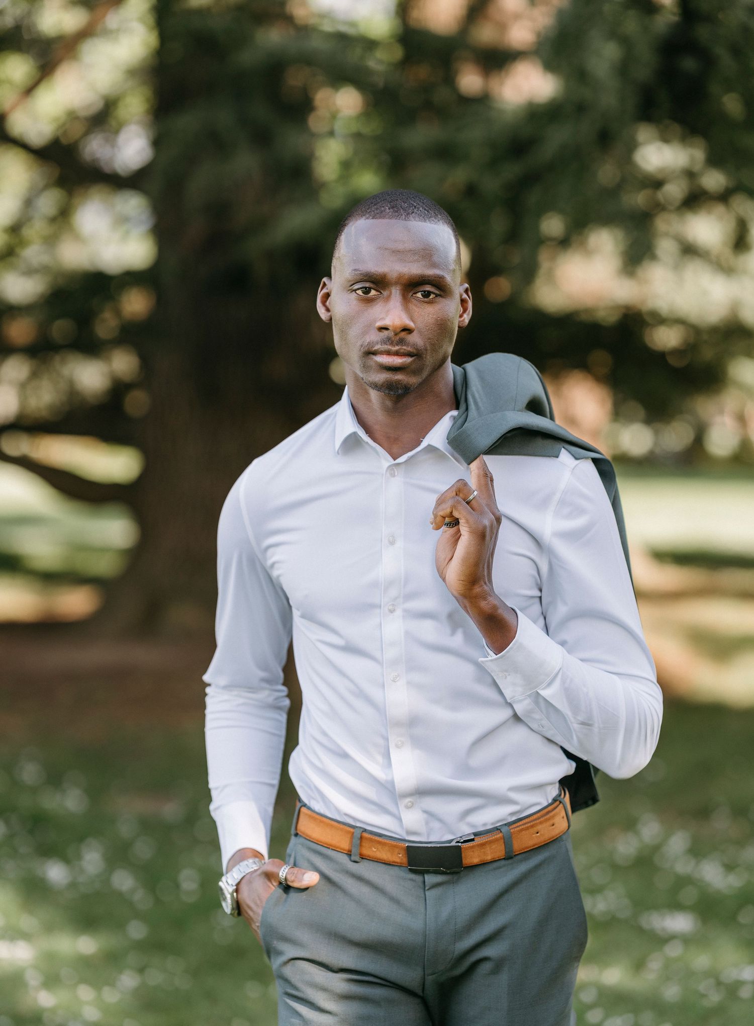 Confident Black man in white shirt carrying suit jacket over shoulder in sunny park.