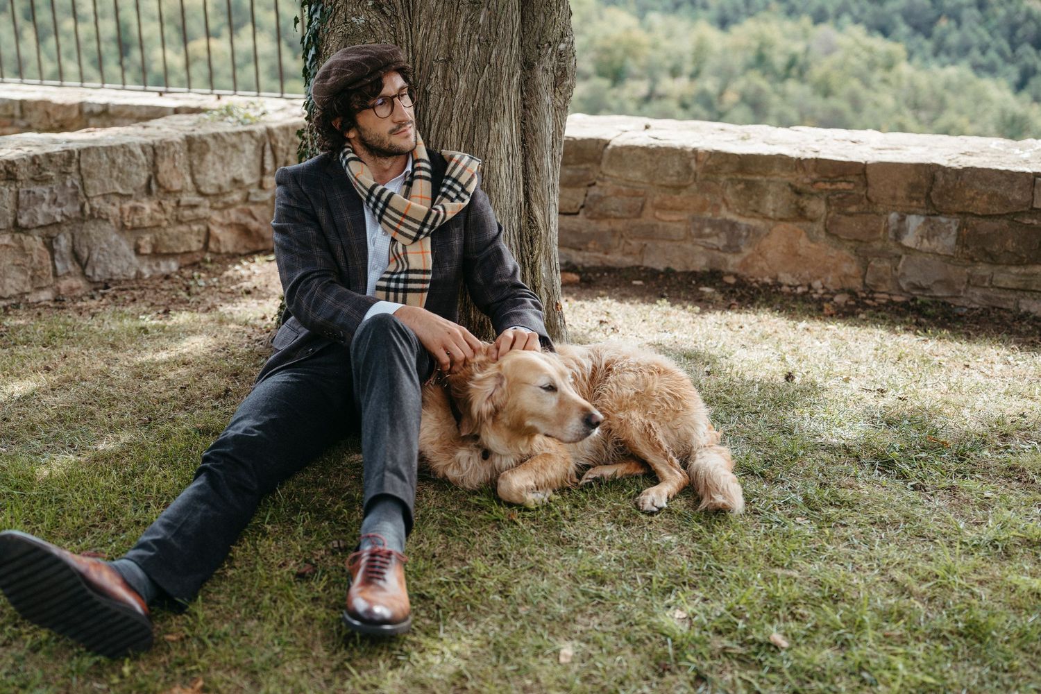 Dapper man in a Burberry scarf sitting on grass under a tree, petting a golden retriever dog.