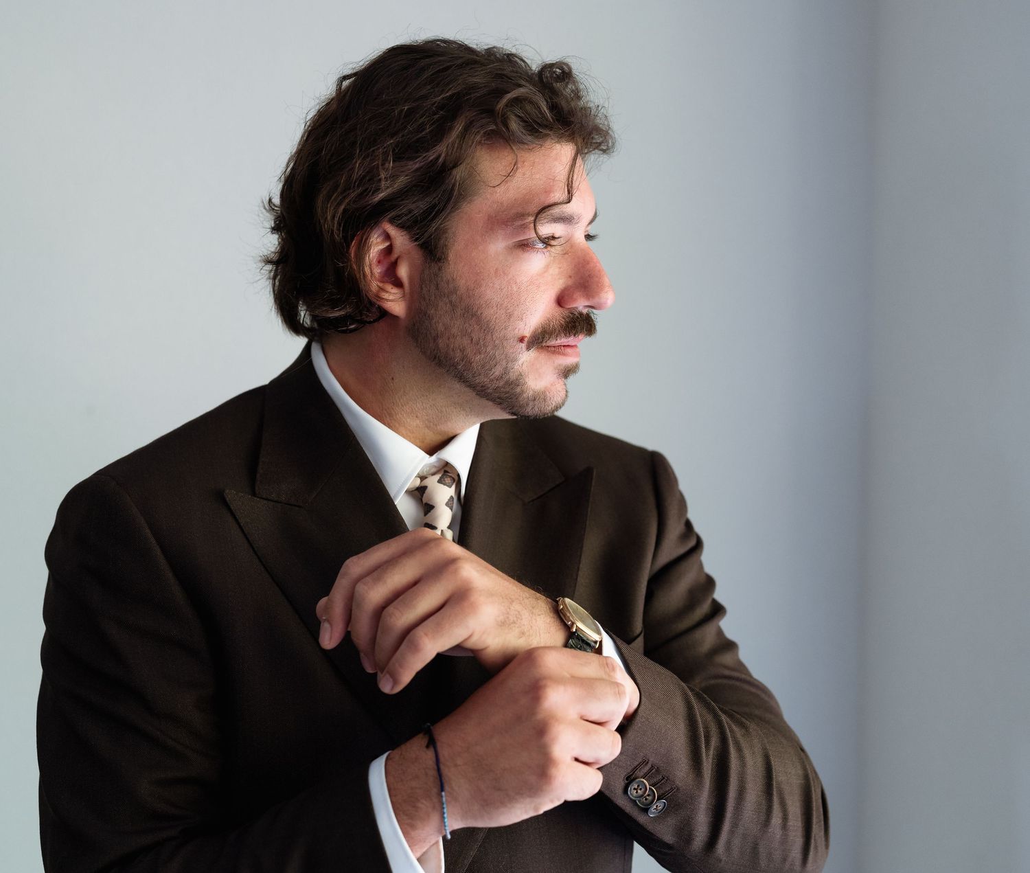 A well-dressed man in a dark suit adjusting his tie, with a mustache and beard, looking sideways against a light background.